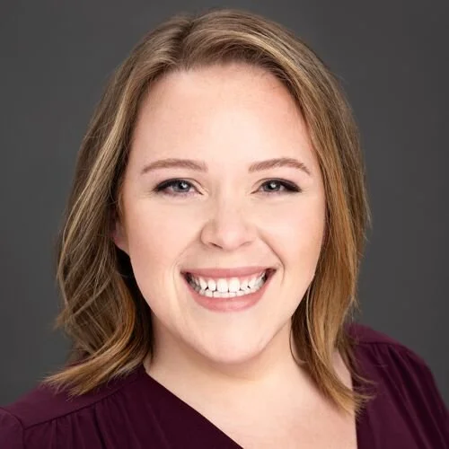 Portrait of a smiling woman with blonde hair and blue eyes, wearing a maroon top and standing against a gray background.