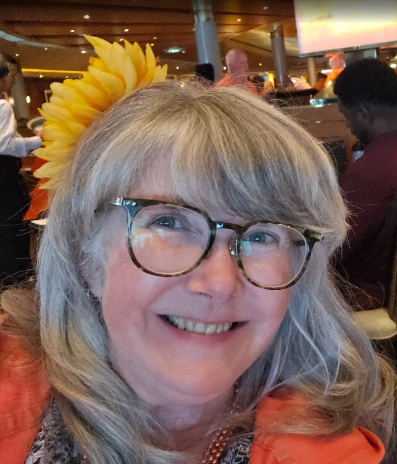 Smiling woman with gray hair and large tortoiseshell glasses, wearing a sunflower hair accessory, in a crowded indoor setting.
