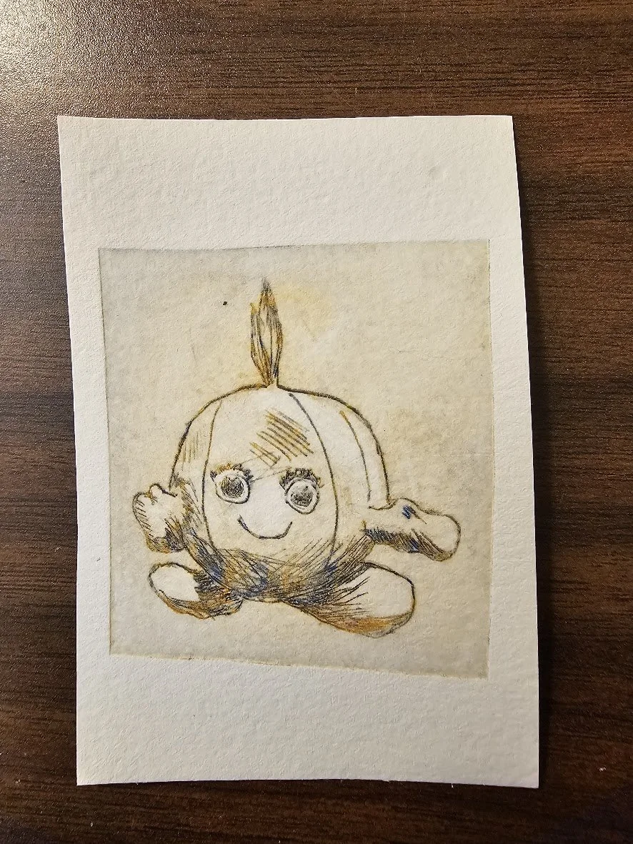 A hand-drawn sketch of a cute round creature with a small tuft of hair on top, large expressive eyes, a smiling mouth, short arms, and big feet, sitting on a piece of paper on a wooden surface.