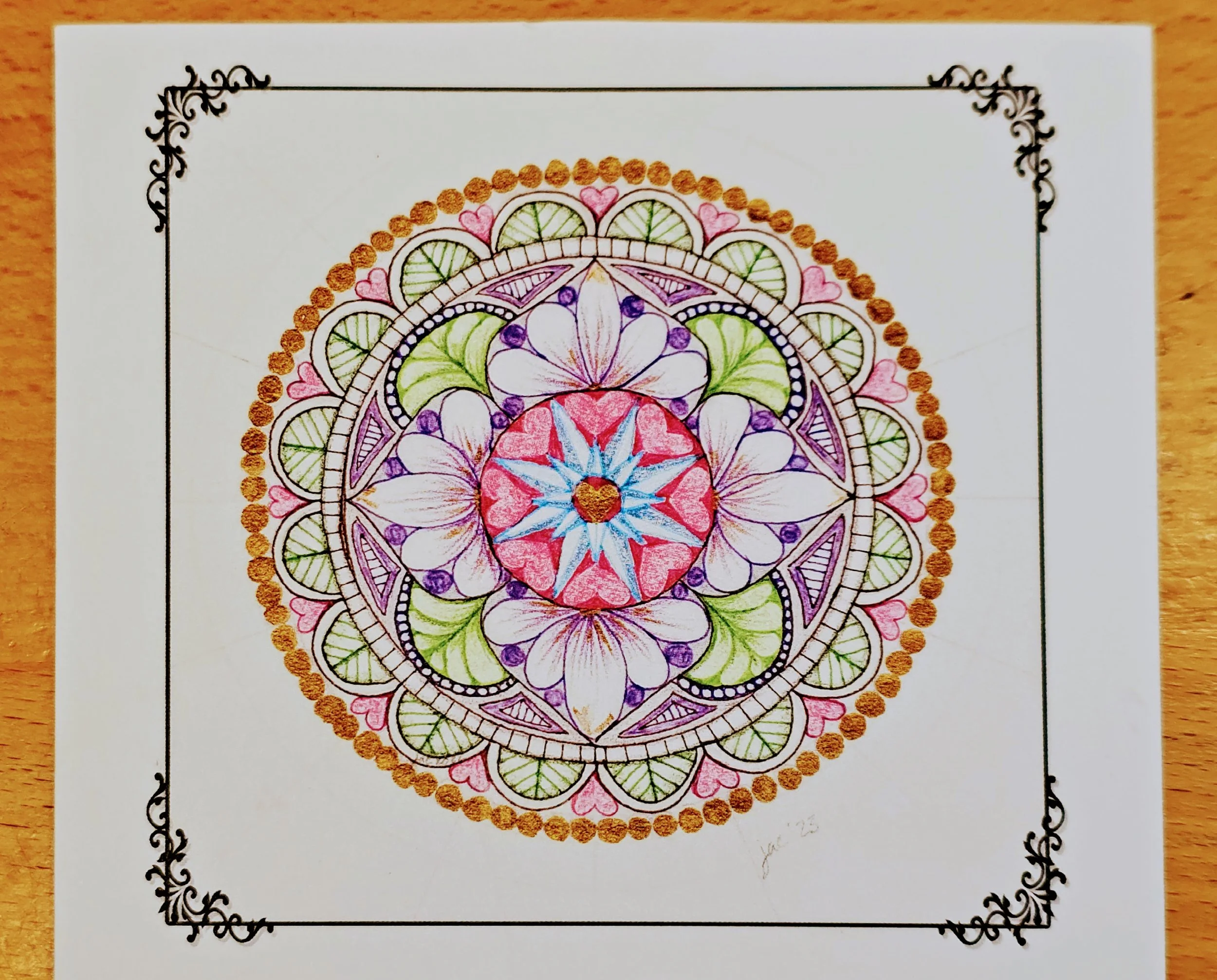 A colorful, intricate mandala drawing on white paper, bordered with decorative black corner designs, placed on a wooden surface.