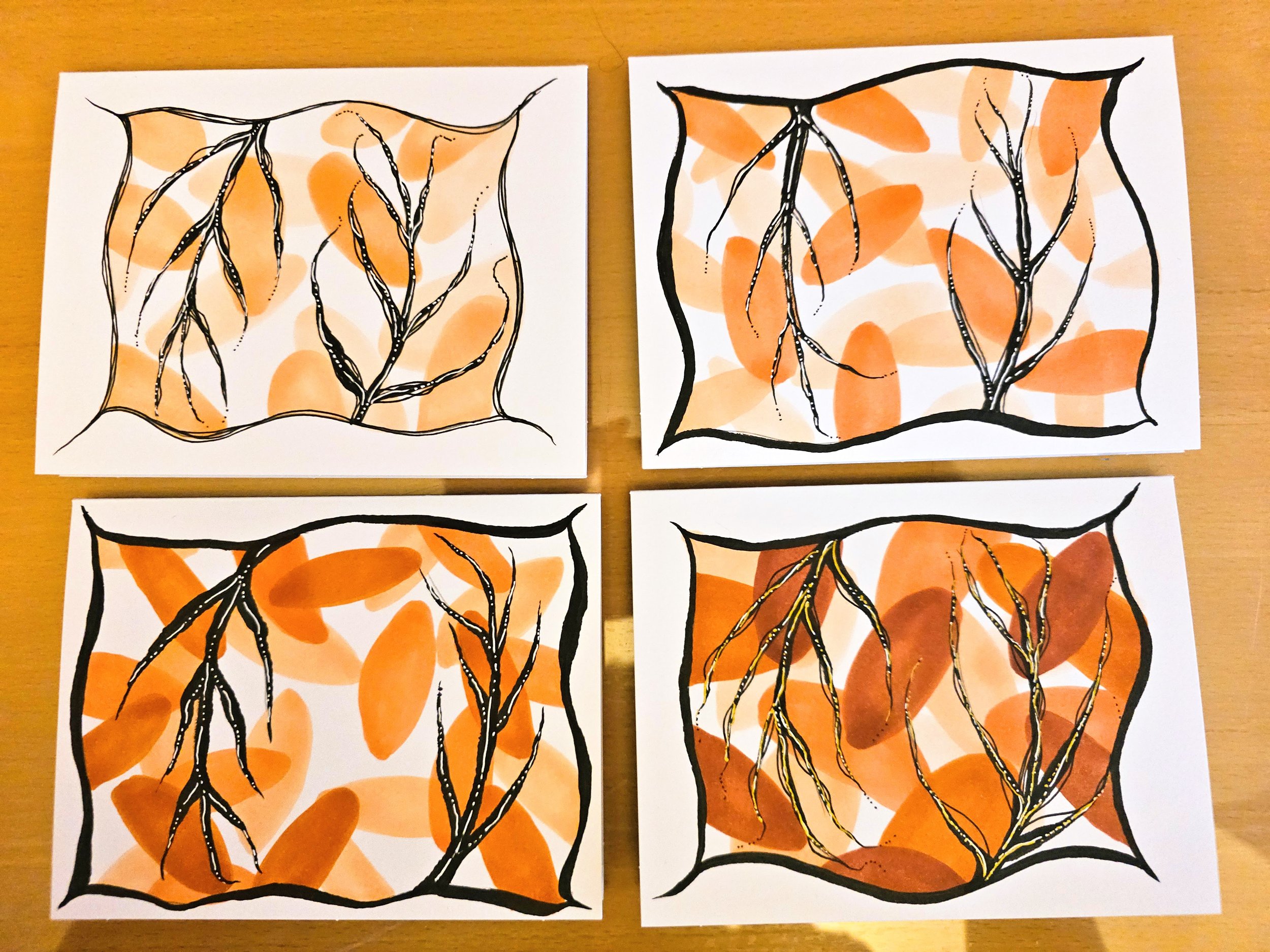 Four handmade art projects on white paper with orange and beige watercolor background, black line drawings of a plant with roots and a border with curved edges displayed on a wooden surface.