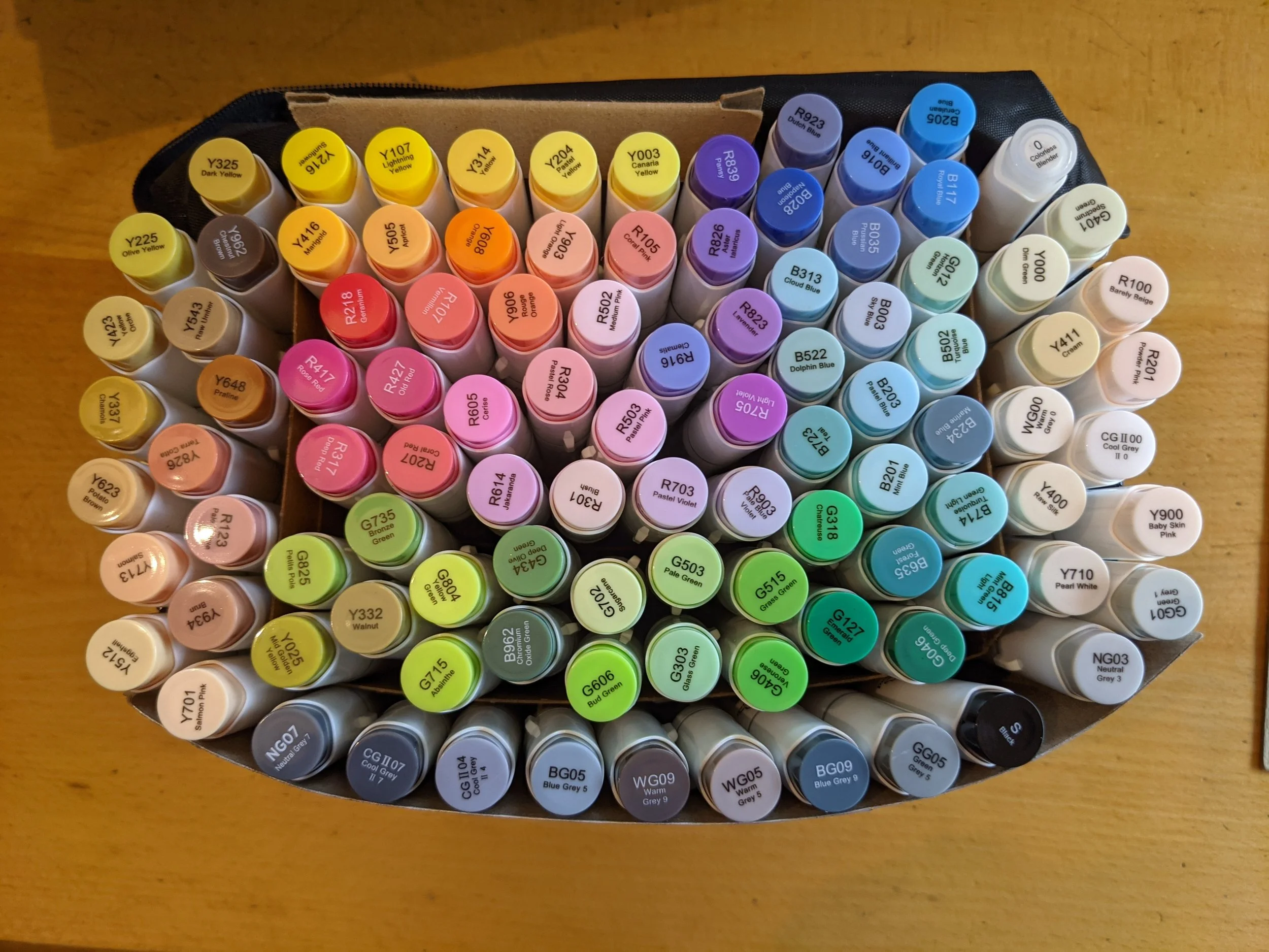 A top-down view of a collection of cylindrical paint marker bottles arranged in a circular pattern on a wooden surface. The bottles are organized by color, with yellow, orange, pink, purple, blue, green, beige, gray, black, and white shades, each labeled with their respective color names and codes.
