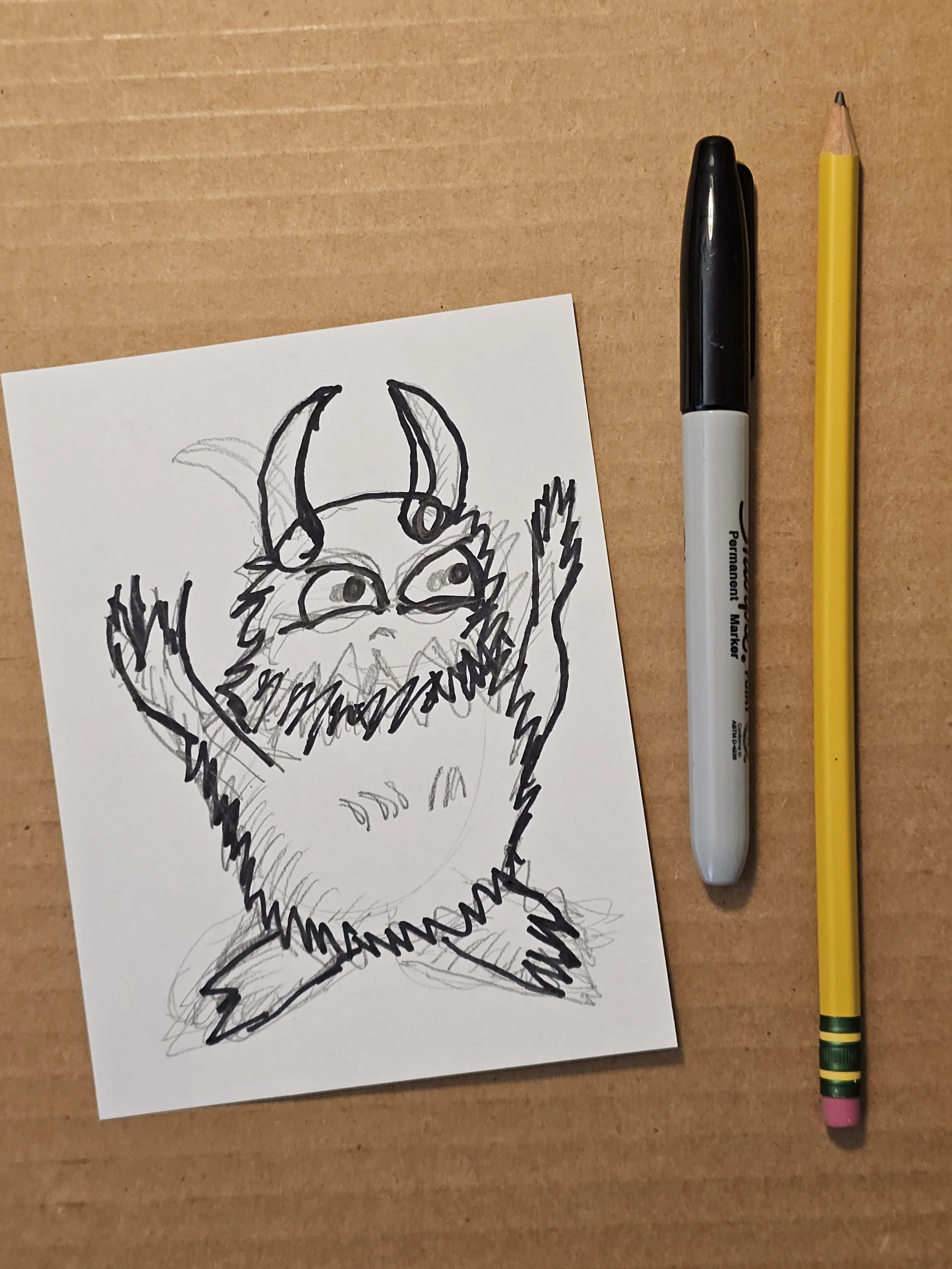A rough sketch of a mythical creature with horns and fur, sketched in black ink on a white piece of paper placed on a wooden surface, accompanied by a black marker and a yellow pencil.