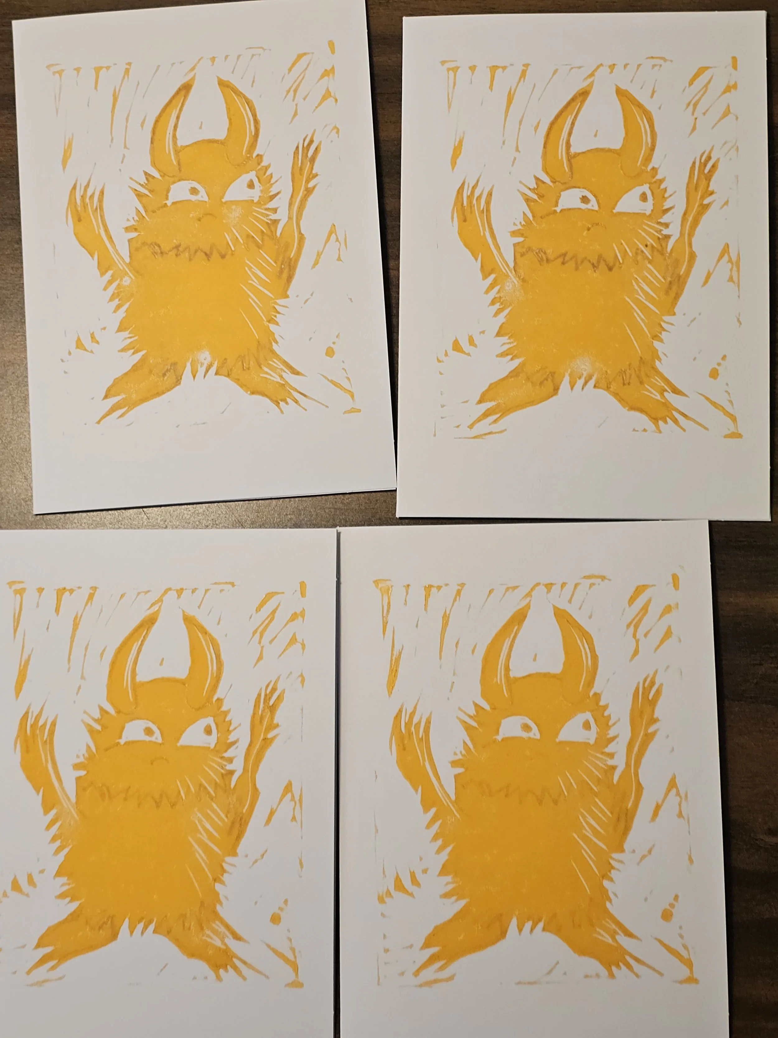 Four sheets of white paper with yellow ink stencil art of a creature with large ears, a beard, and a smiling face, arranged in a two-by-two grid on a dark wooden surface.