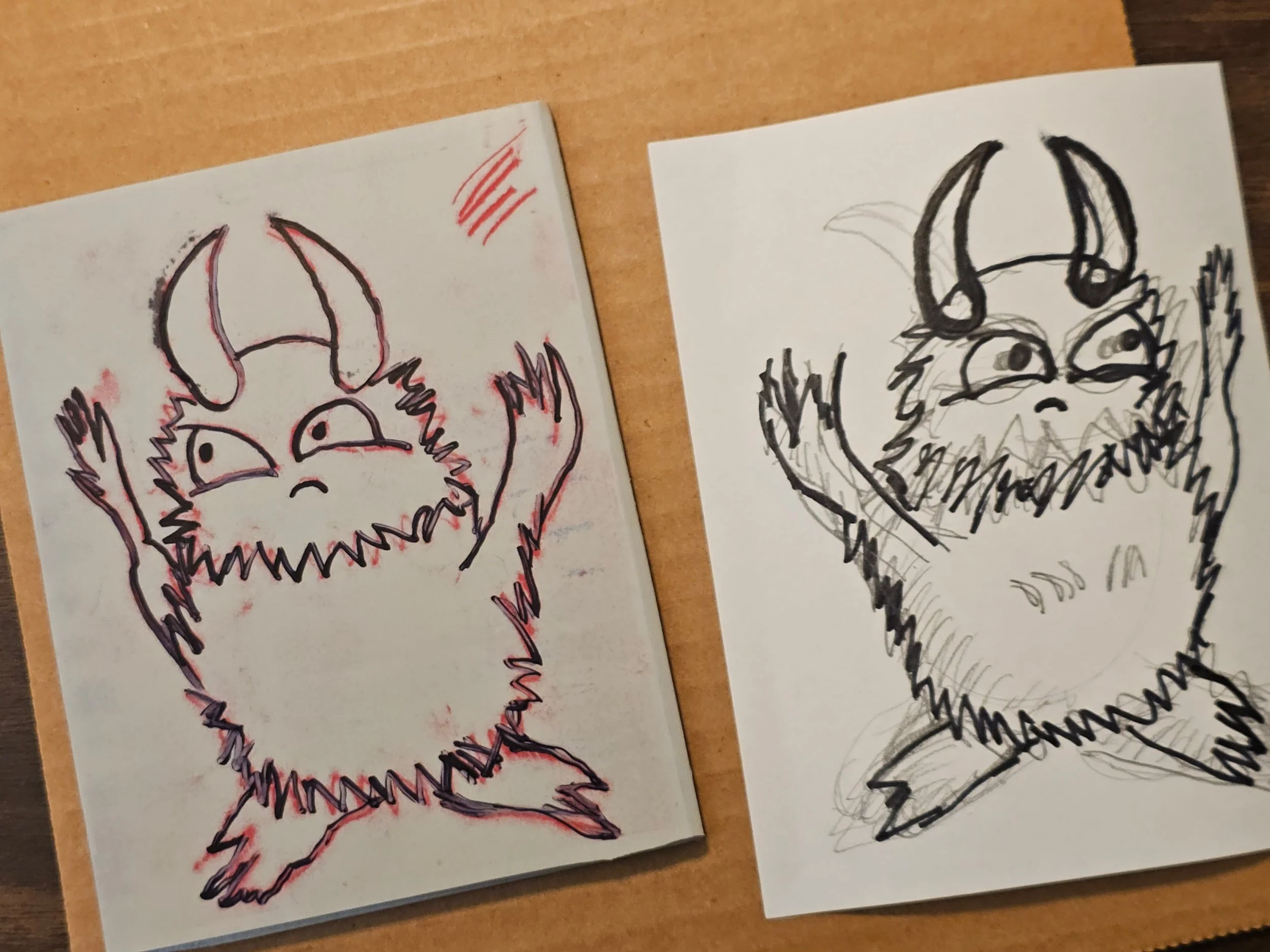 Two pieces of paper, each with a sketch of a character's face, on a wooden surface. The left sketch is drawn with red and black ink, showing a creature with horns and sharp teeth. The right sketch is black ink, depicting a similar character with detailed features.