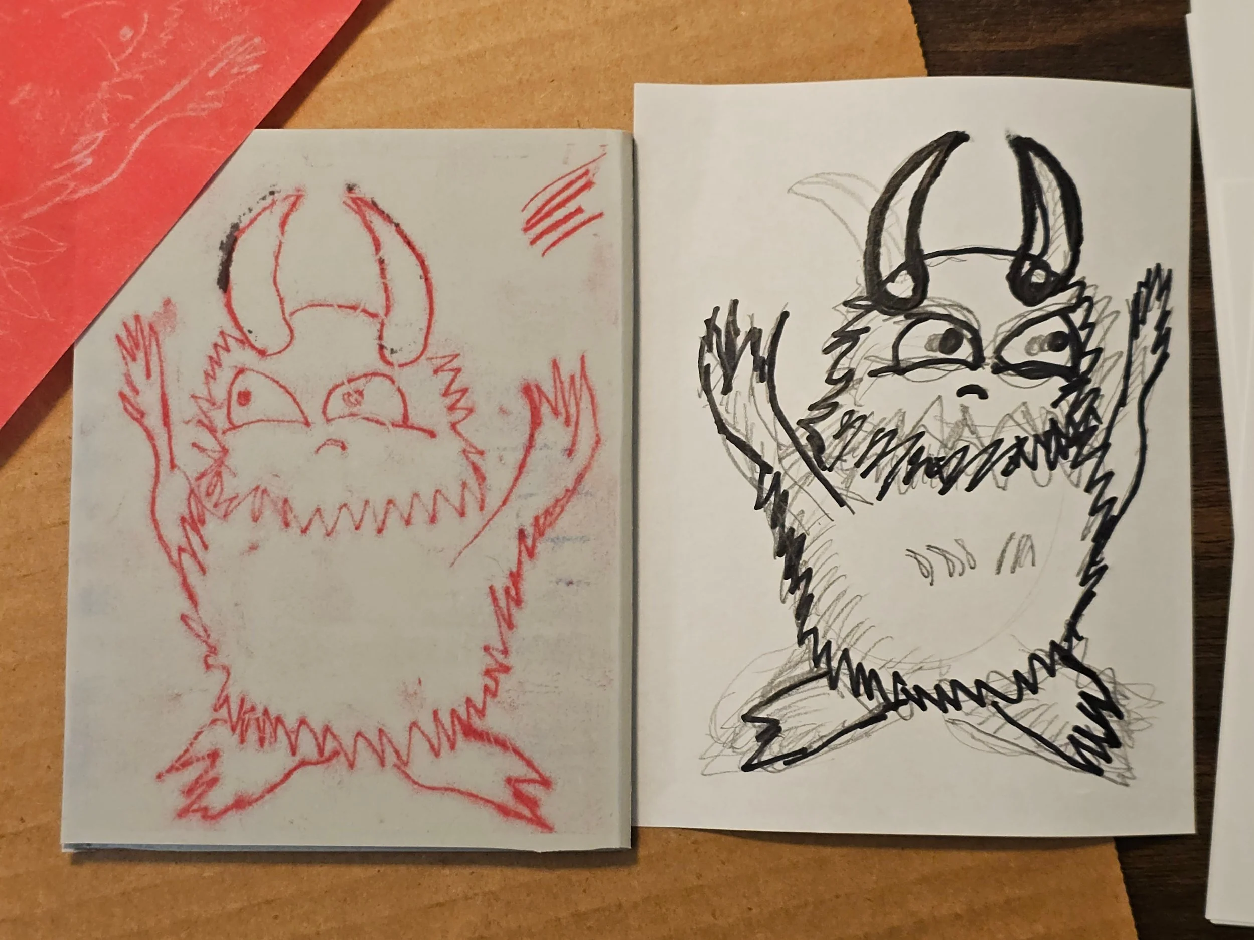 Two sketches of a fox-like creature, one in red ink and the other in black ink, on separate pieces of paper placed on a wooden surface.