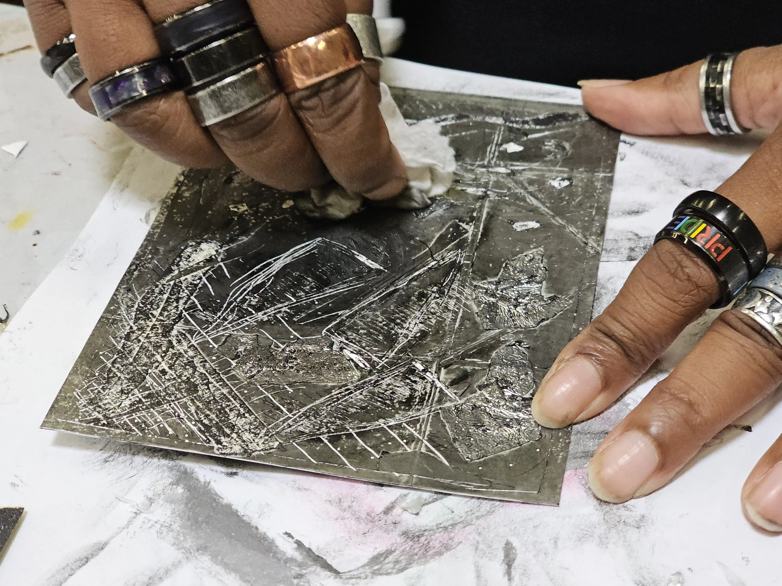 Close-up of a person's hands wearing multiple rings and a black wristband with rainbow-colored text, working with a piece of shiny silver metal, possibly engraving or carving, on a gray work surface.
