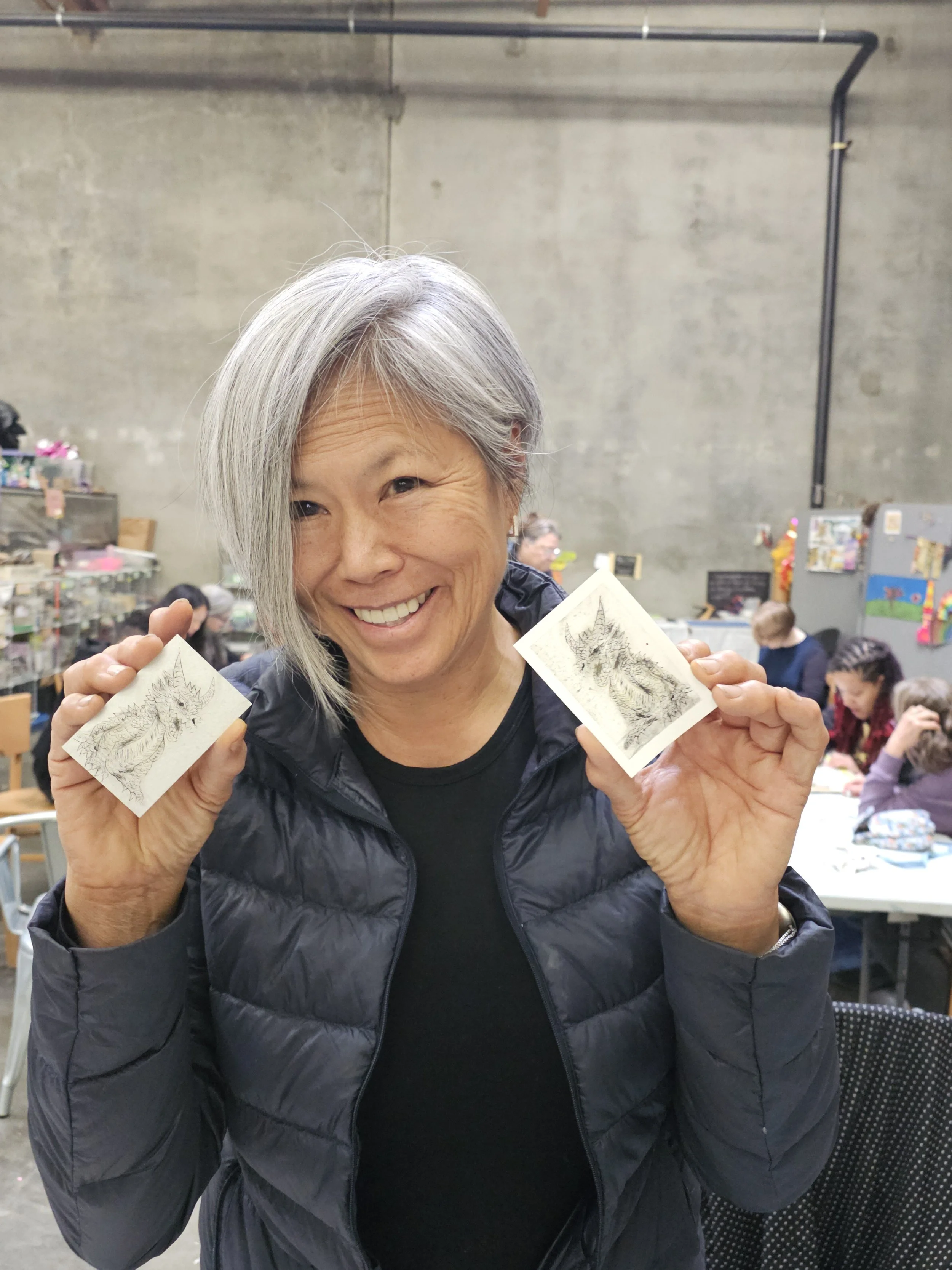 A woman with short gray hair smiling and holding two illustrated cards of a mythical creature, a dragon, in a busy indoor marketplace or craft fair setting.