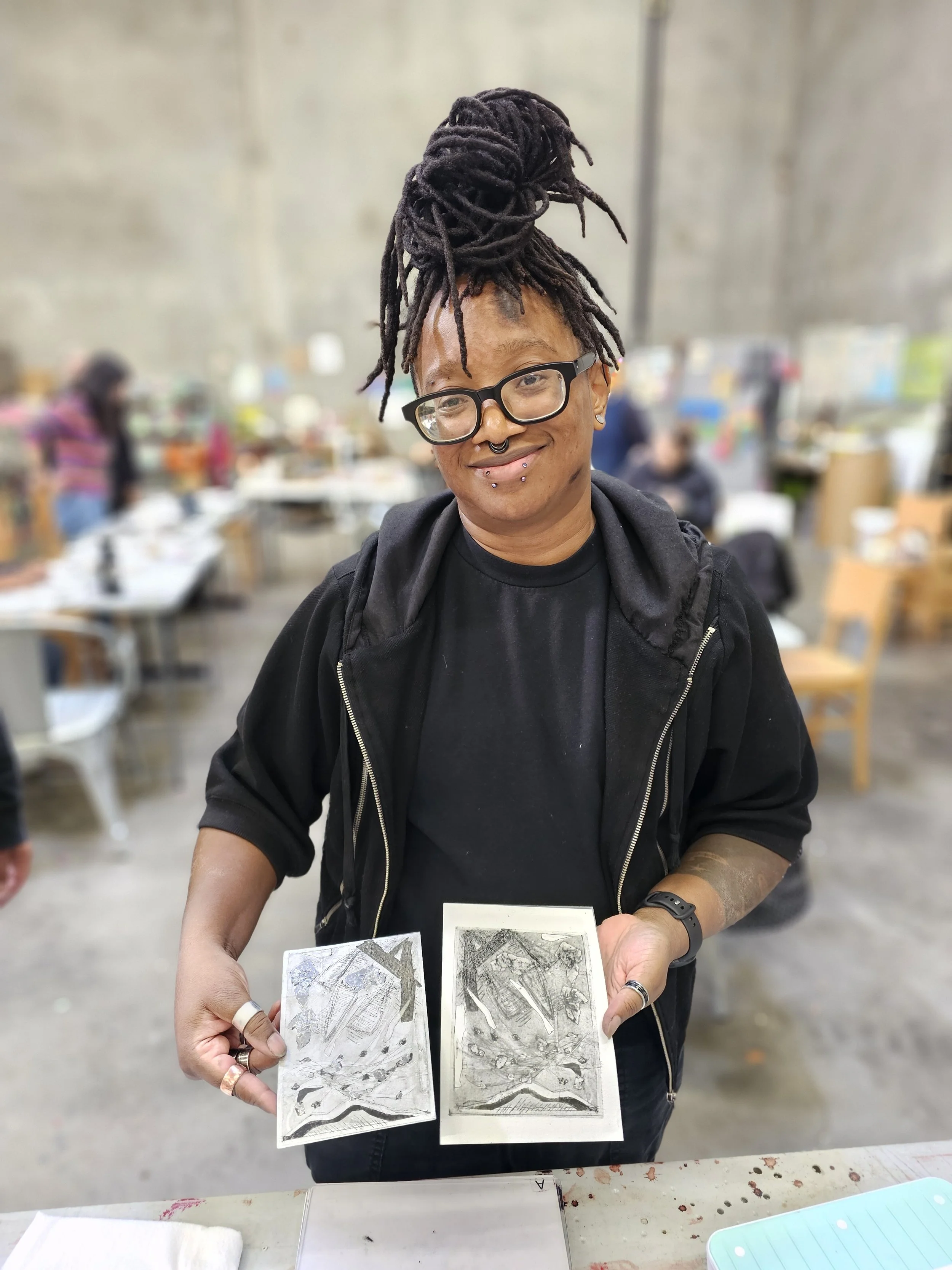 Person with dreadlocks and glasses holding black and white abstract artwork in an indoor setting.