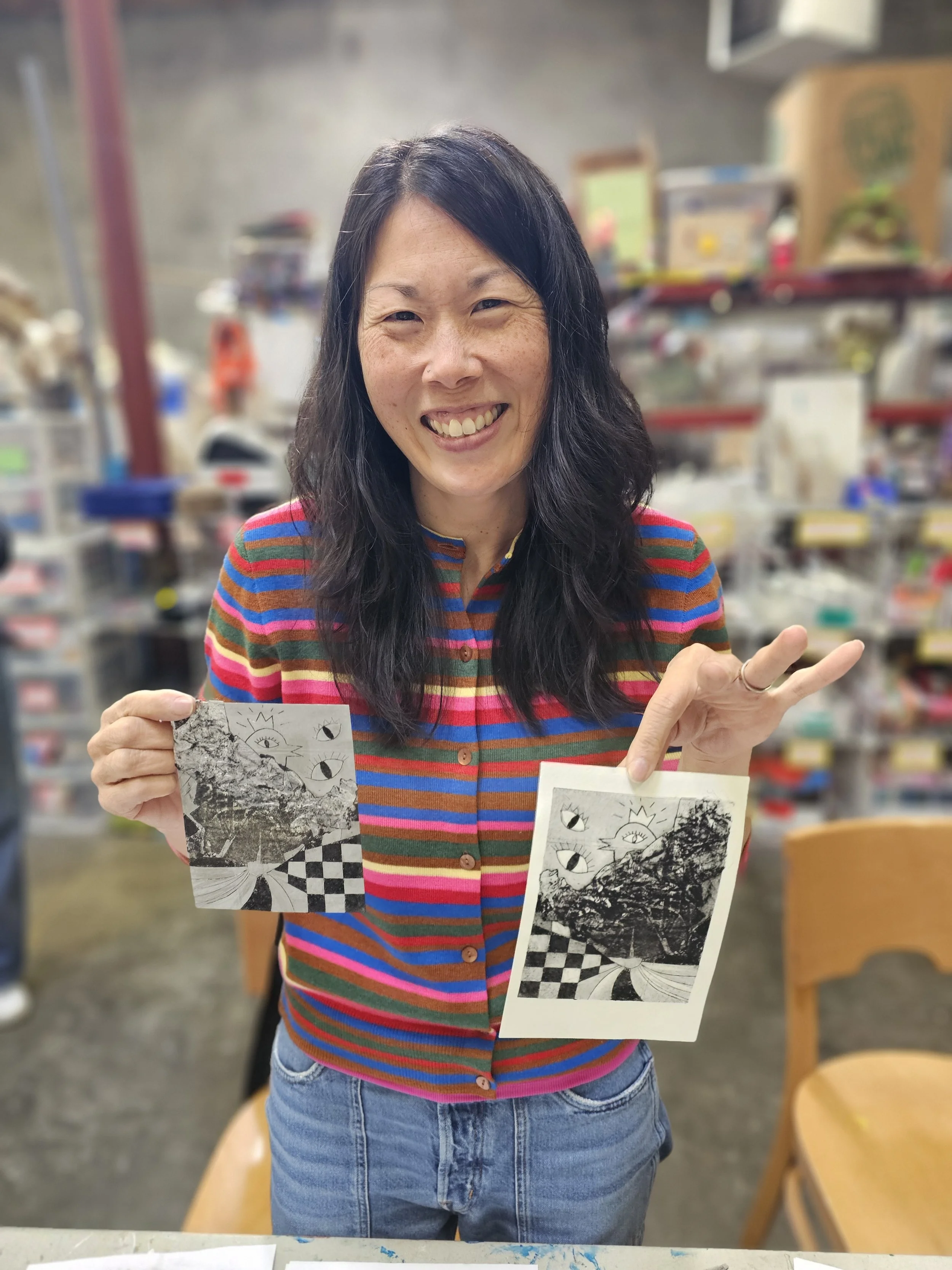 A woman holding black and white drawings in a room with shelves and various supplies, smiling and making a gesture with her hand.