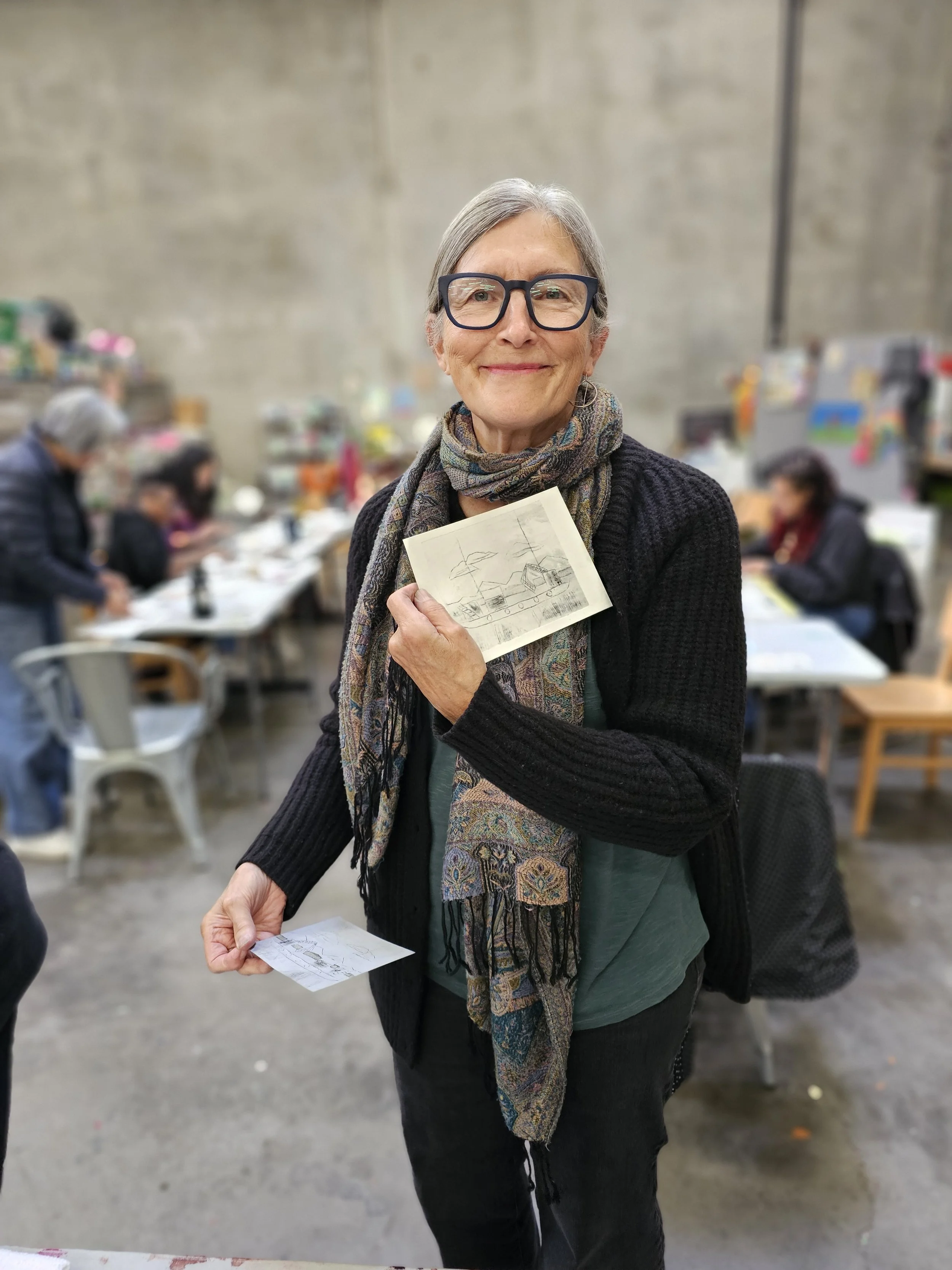 An older woman with glasses and gray hair smiling while holding a sketch of a house, with people sitting and working at tables in the background.
