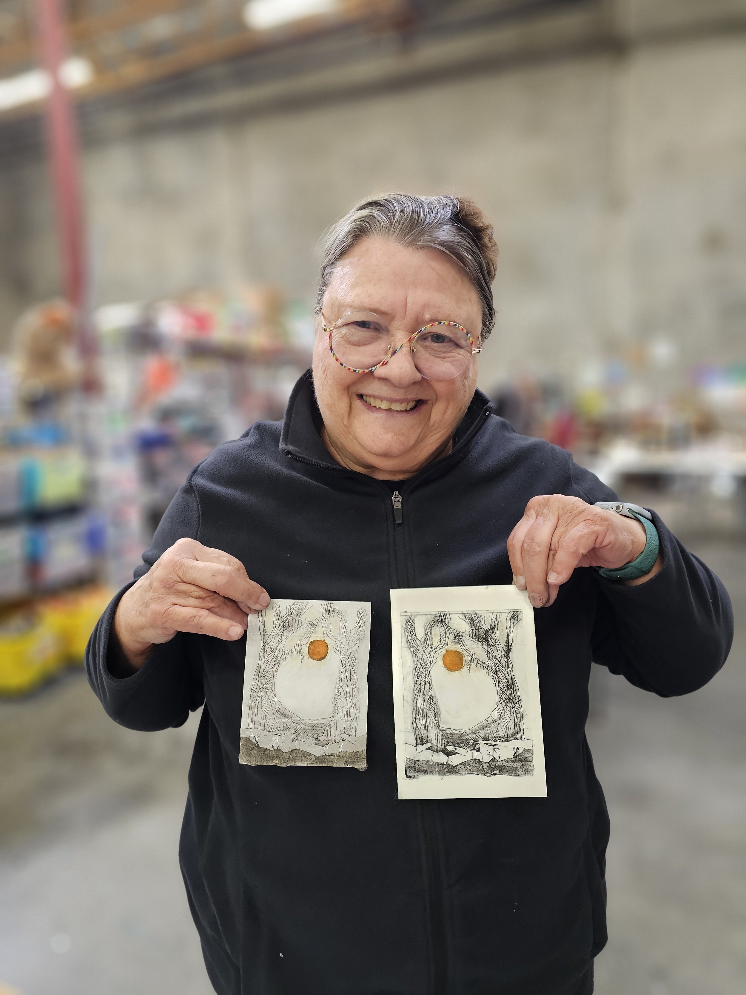 An elderly woman smiling, holding up two small pieces of artwork with a central orange circle and textured background, in a store or warehouse setting.