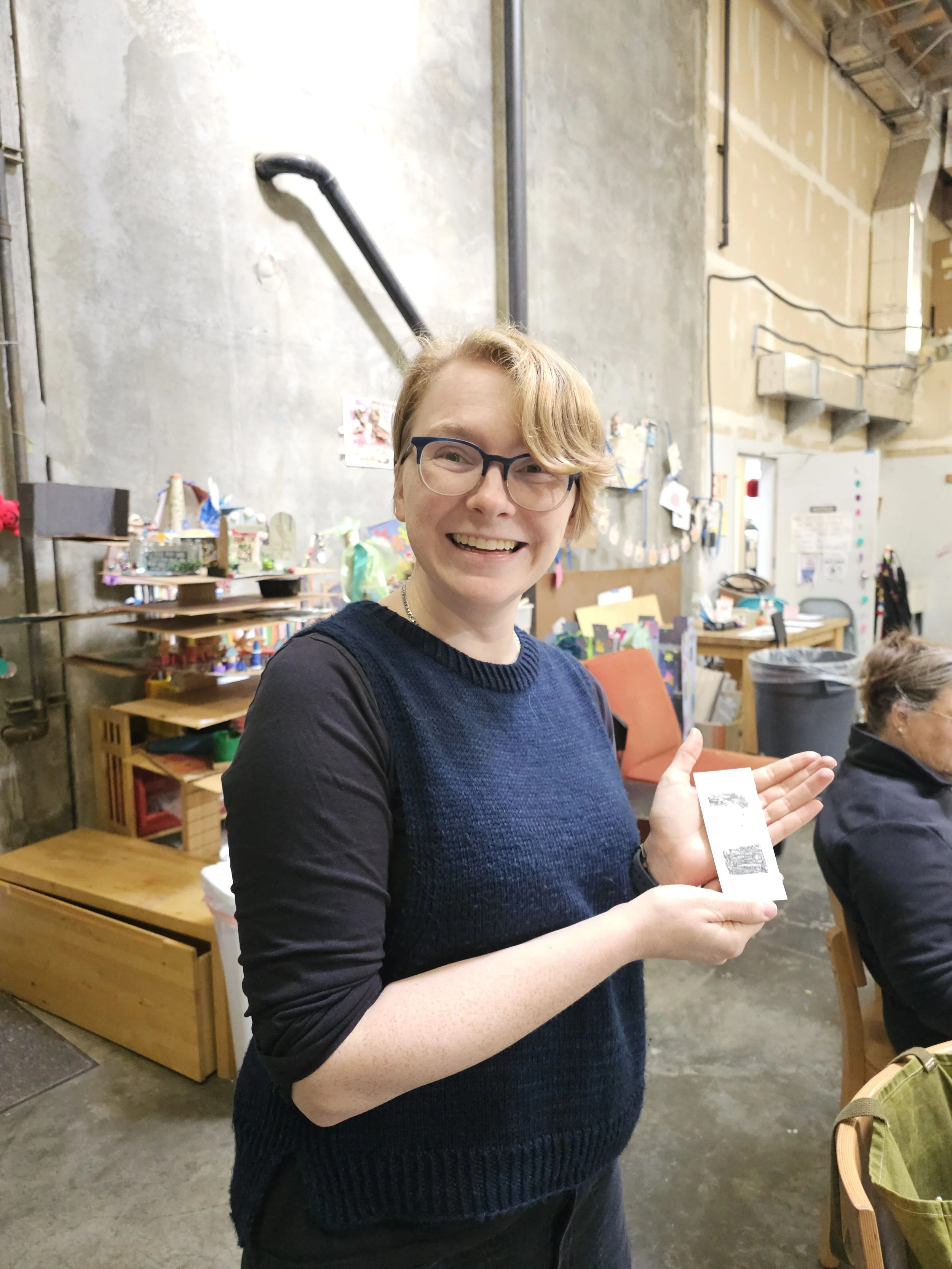A woman with glasses, short blonde hair, wearing a black shirt and blue sweater, smiling and holding a piece of paper or ticket in an indoor, workshop-like setting.