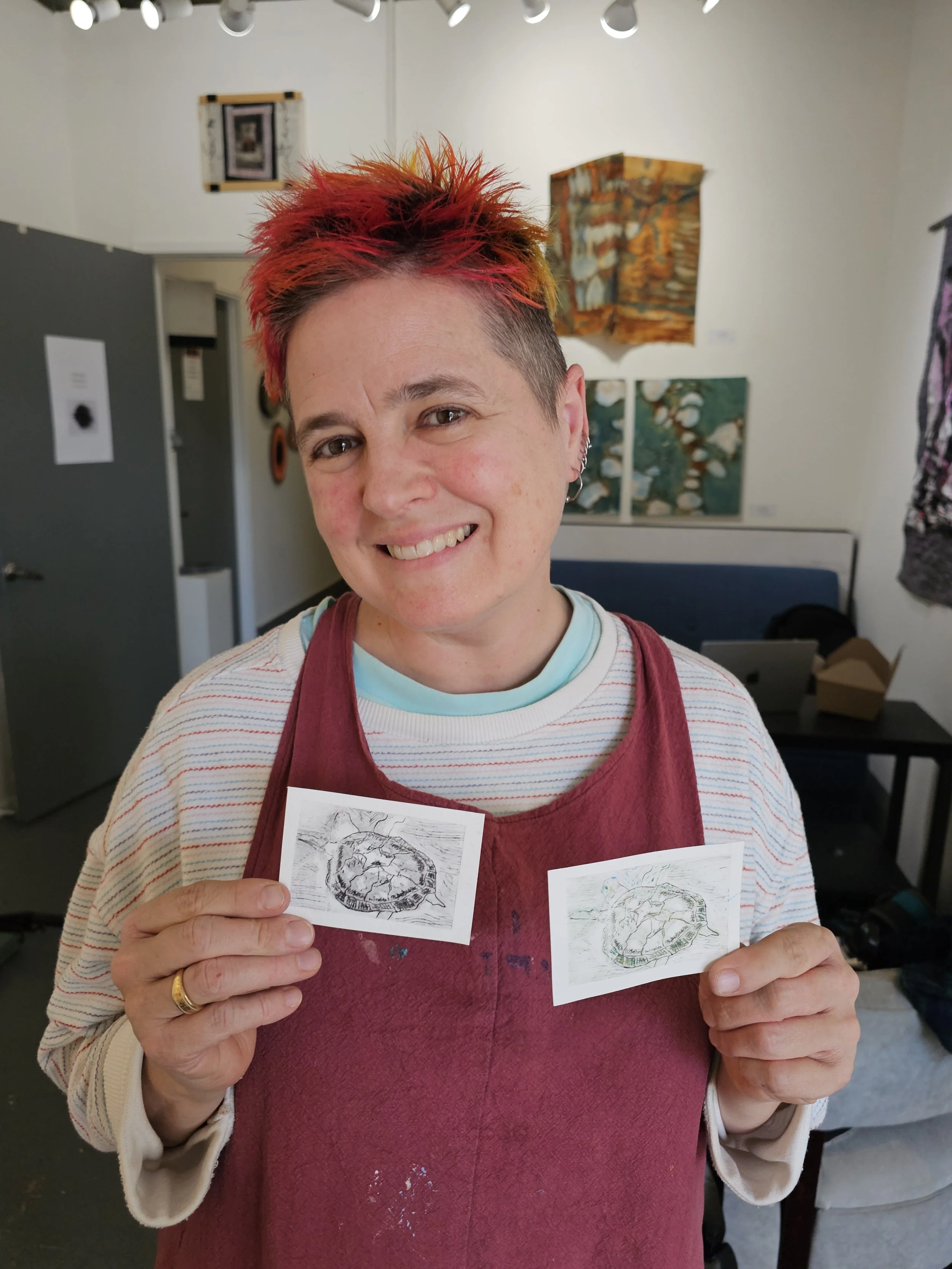 A person with short, spiky hair painted in bright red, holding three small art prints featuring sketches of a lion. The person is smiling and in an art gallery or studio with paintings on the wall and art supplies visible in the background.
