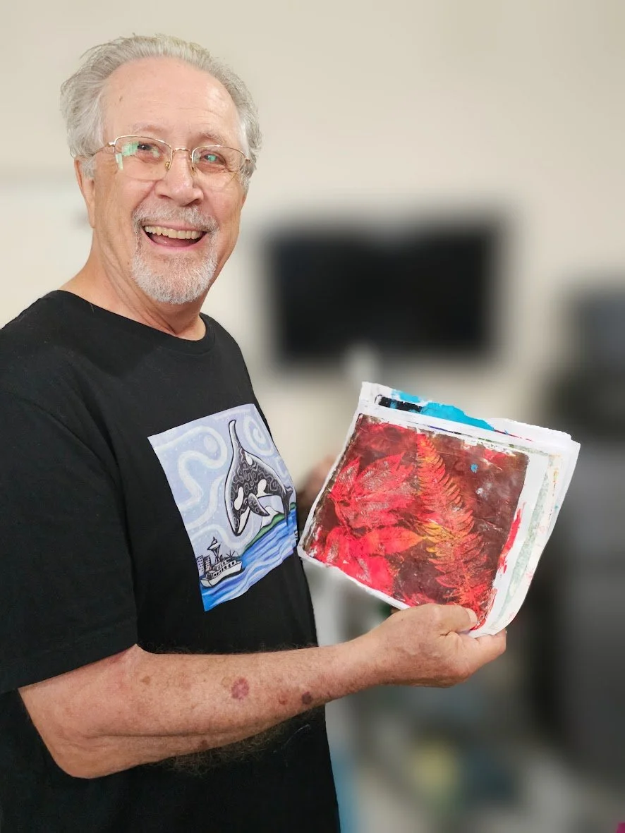 An older man with glasses and gray hair holding a stack of colorful painted papers, smiling at the camera.