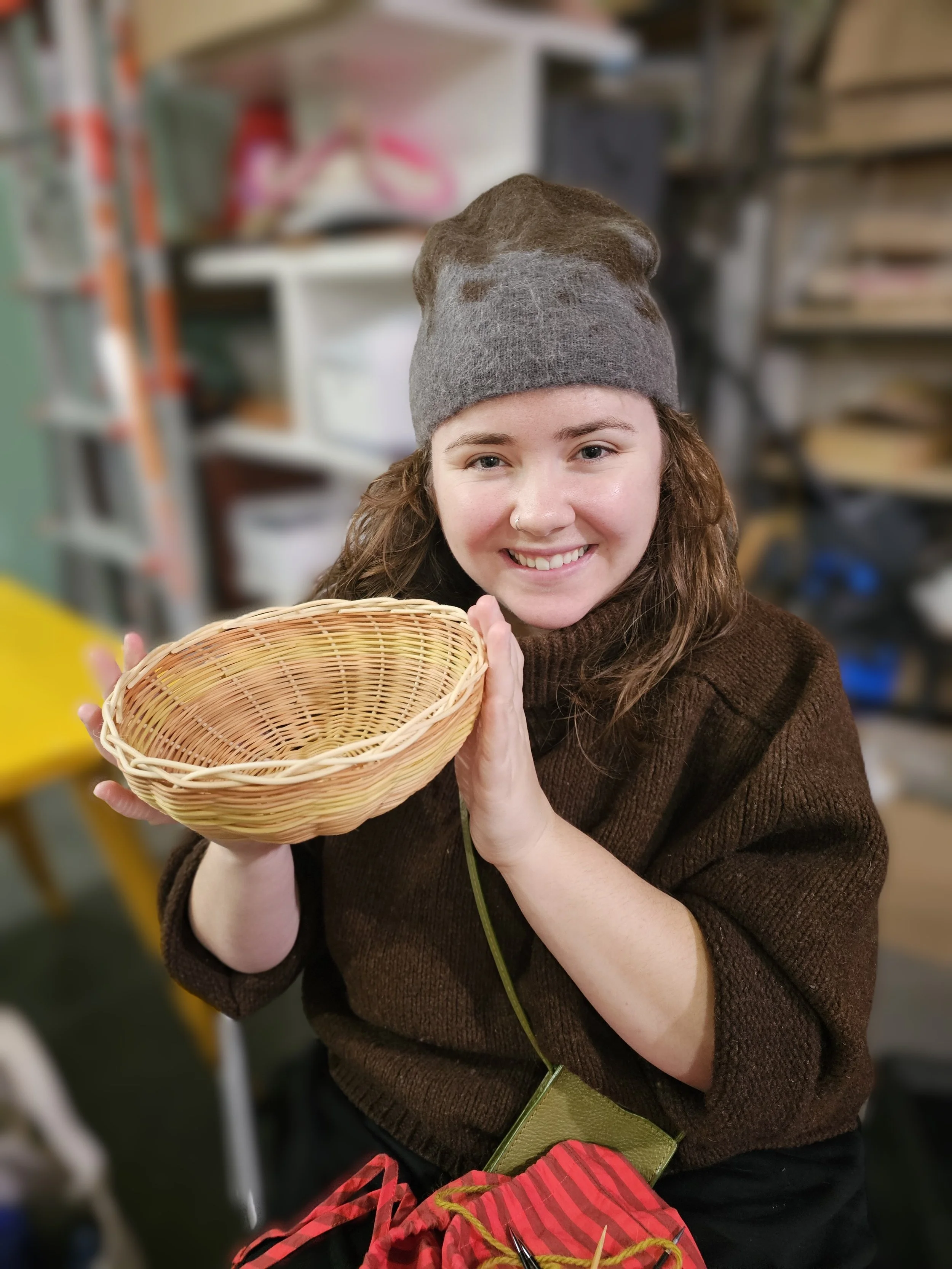 Smiling woman in a gray knit beanie holding an empty woven basket in a craft store