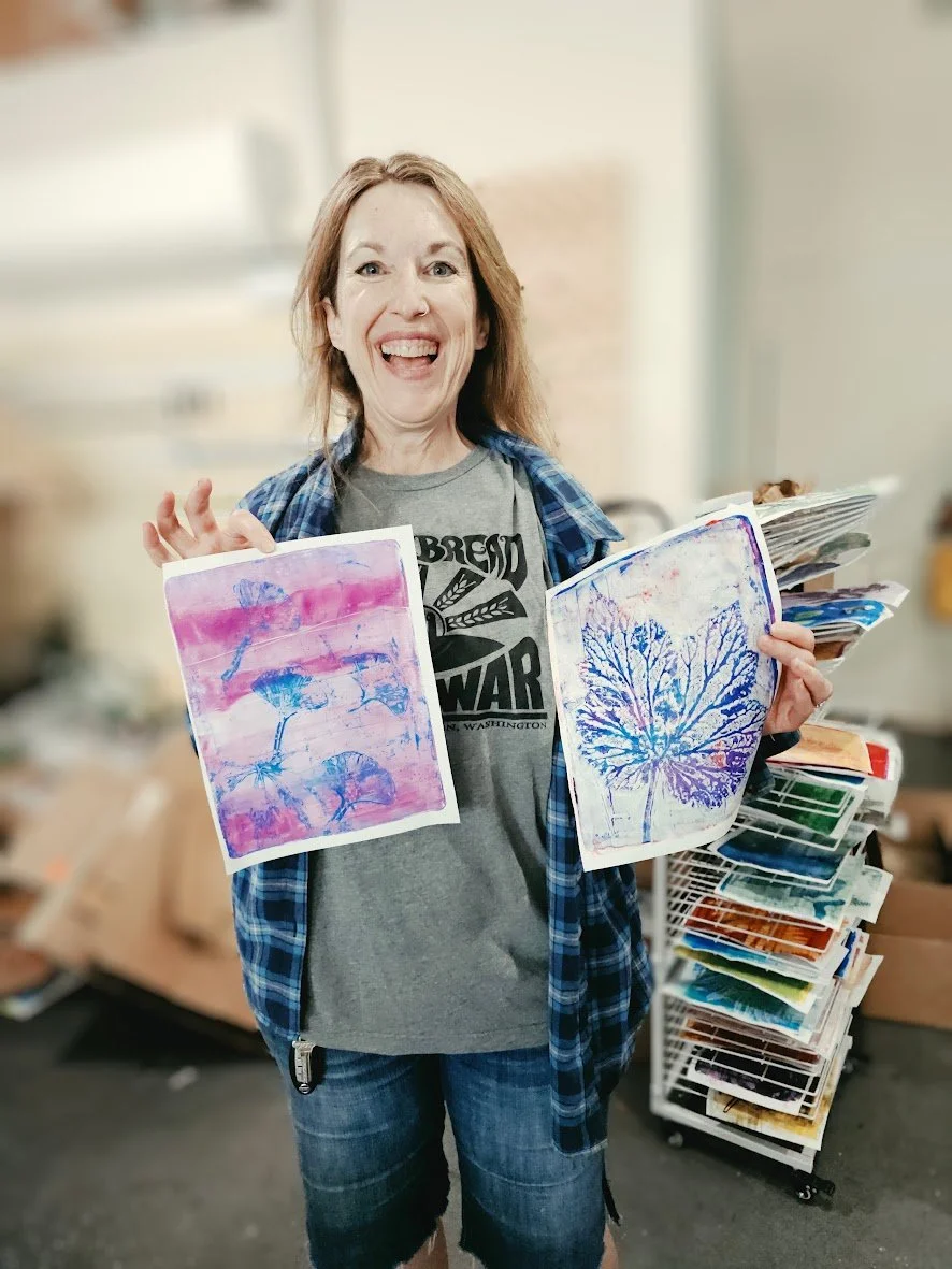 A woman with red hair smiling and holding up two colorful prints of tree silhouettes, standing in an art studio with stacks of artwork and art supplies in the background.