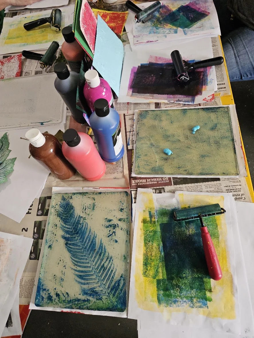 Art supplies on a table, including paint bottles in various colors, rollers, stenciled papers, and paper with painted and stamped textures.