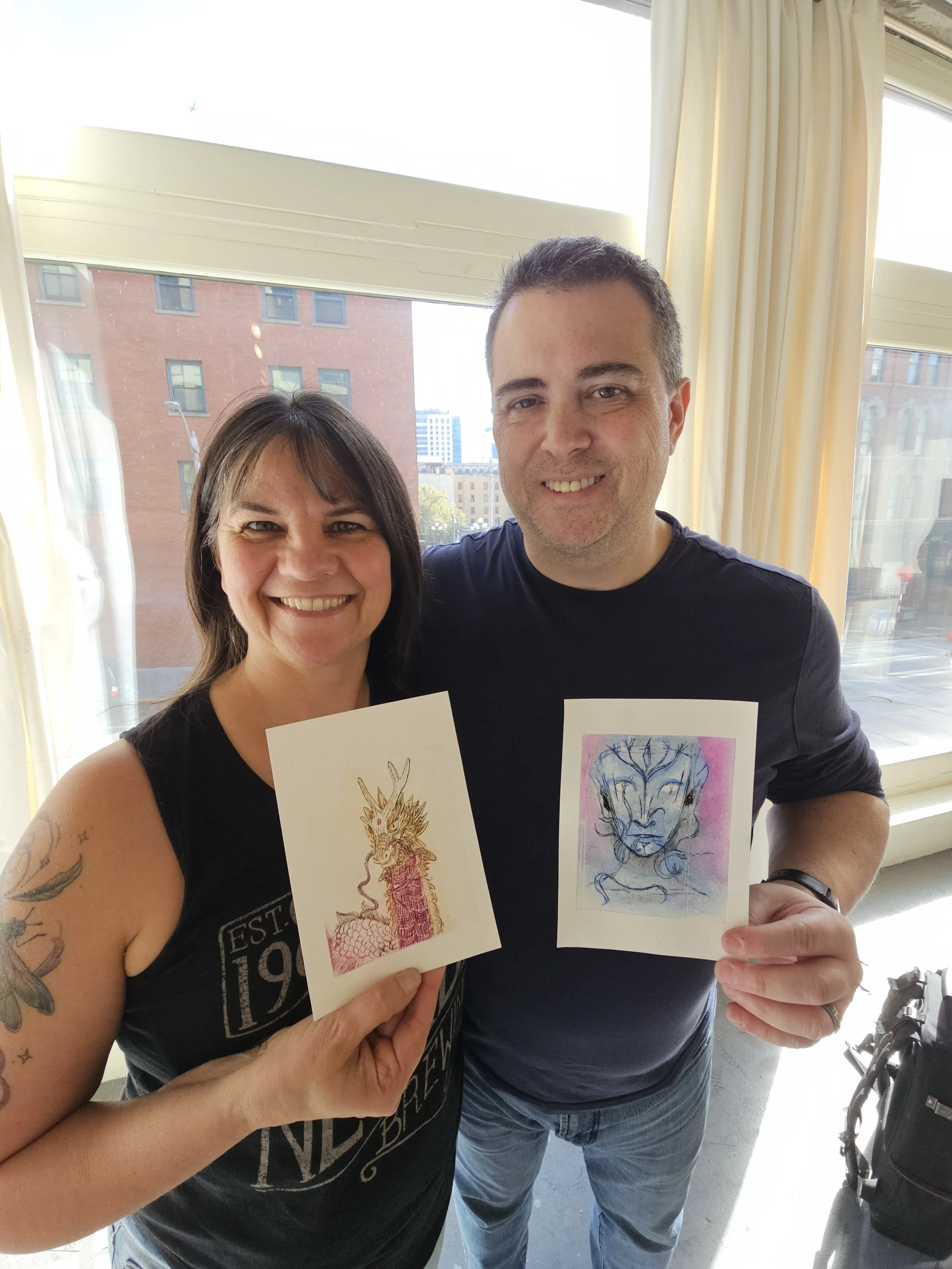 A woman and a man smiling and holding two art prints of fantasy creature designs in front of a window with cityscape outside.