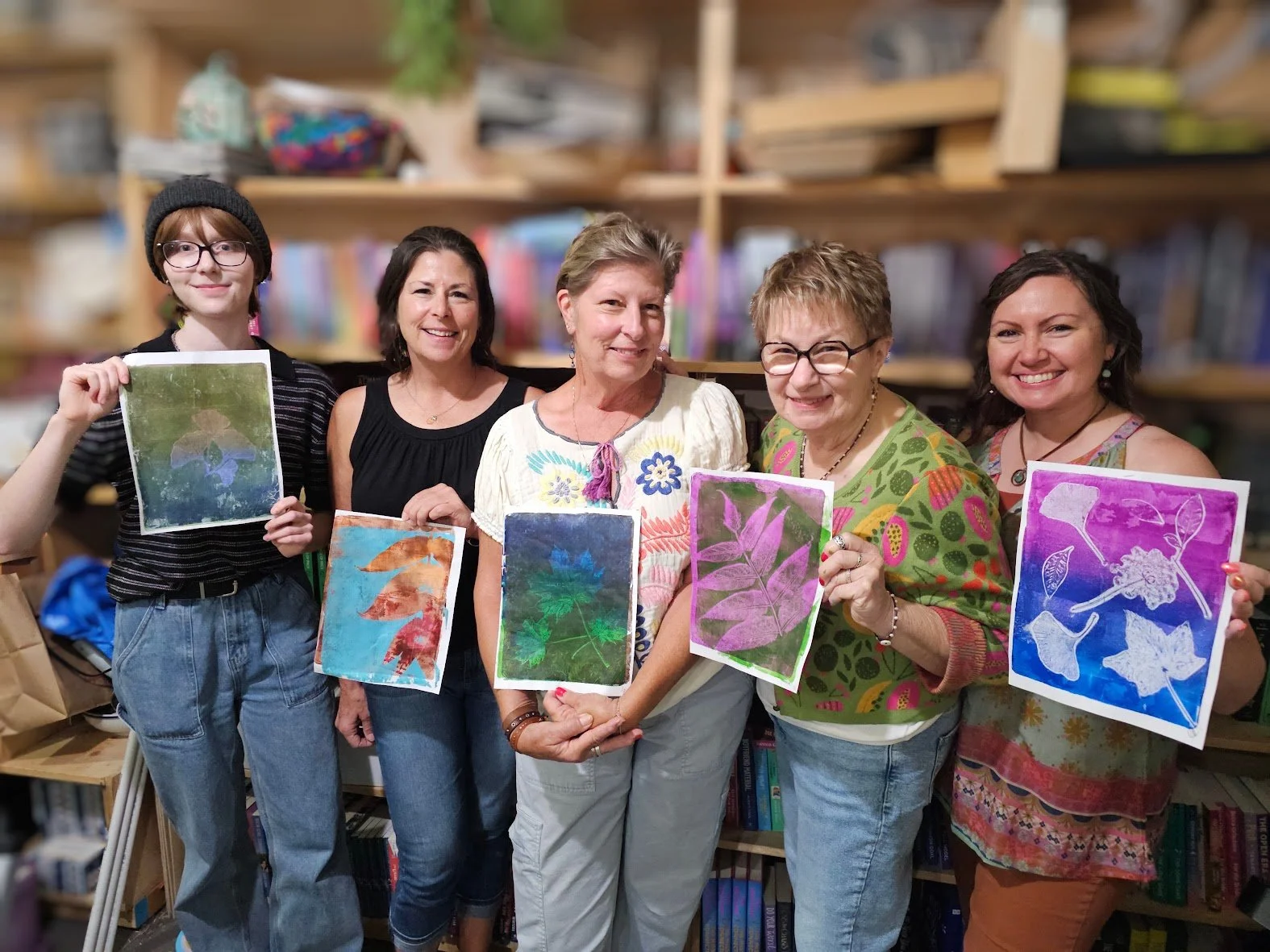 Group of six women and a young girl smiling and holding colorful leaf prints in a library or classroom setting.