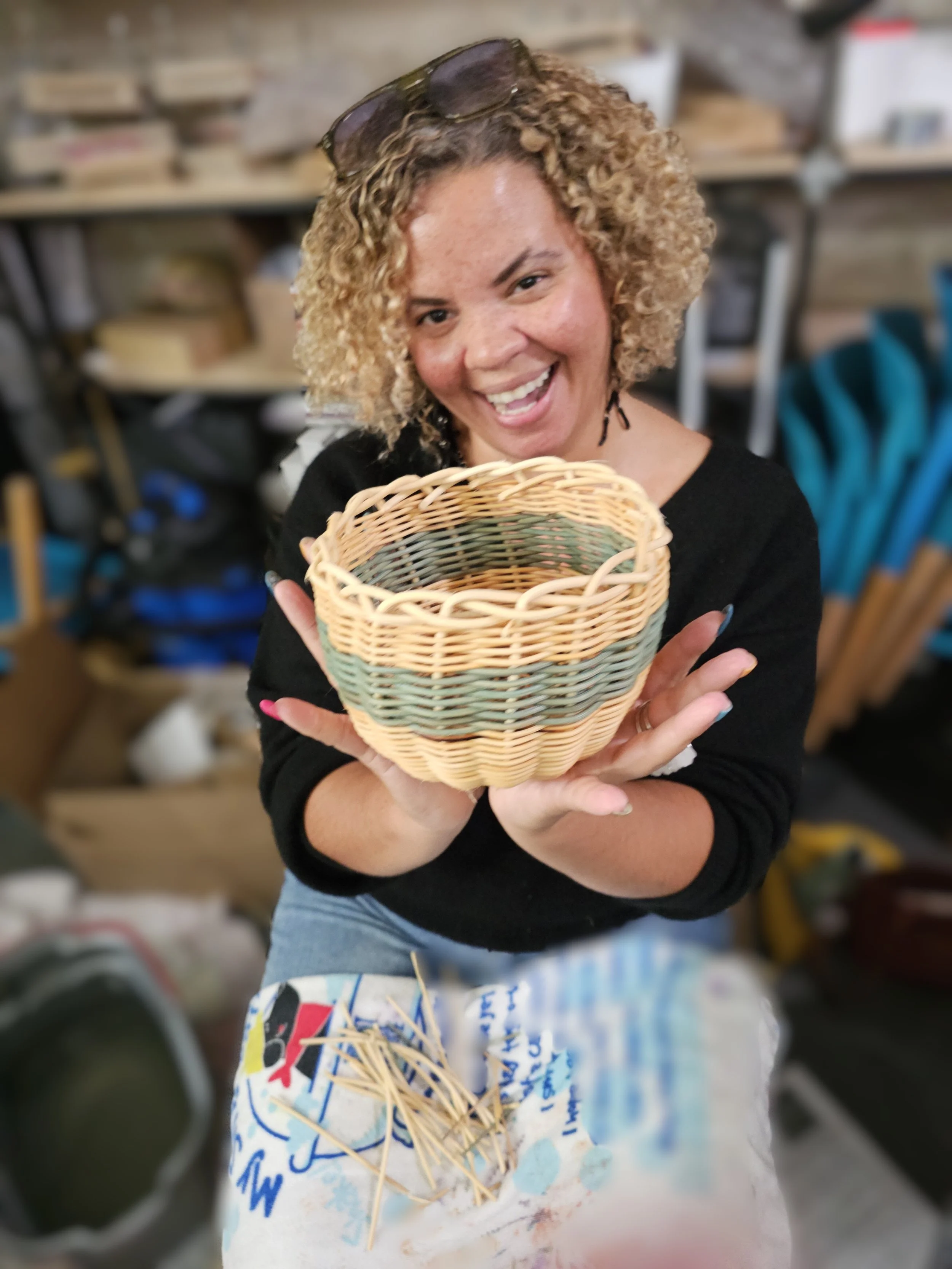 Smiling woman with curly hair holding a handmade woven basket in a craft workshop