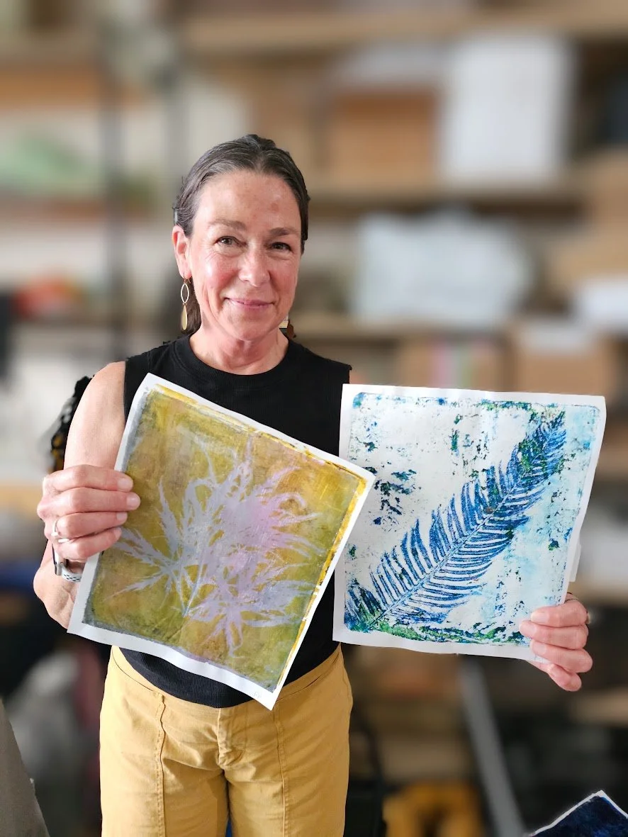 A woman smiling and holding two pieces of artwork with plant prints, standing in an art studio with shelves in the background.