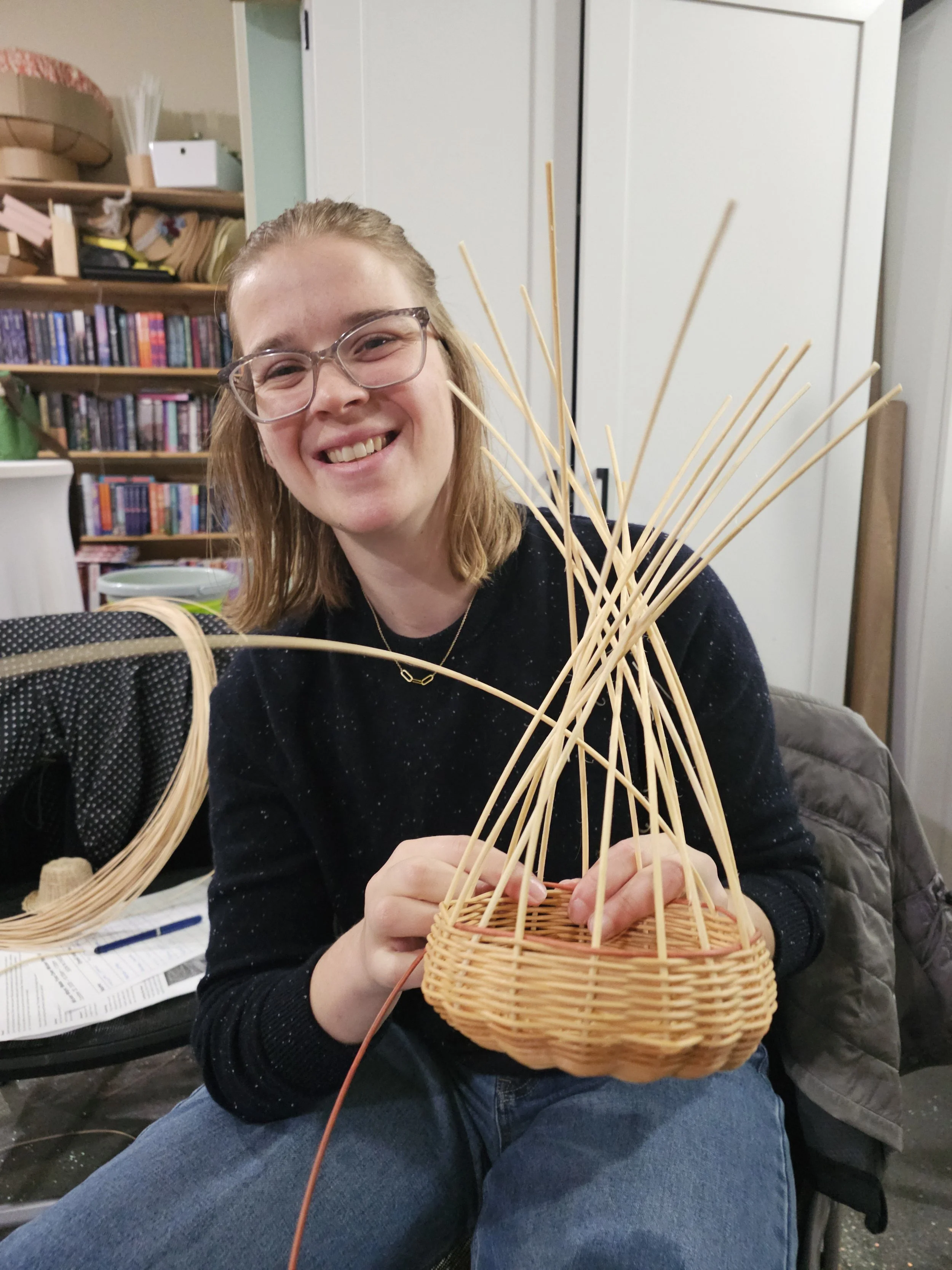 Smiling woman wearing glasses weaves a wicker basket at home.