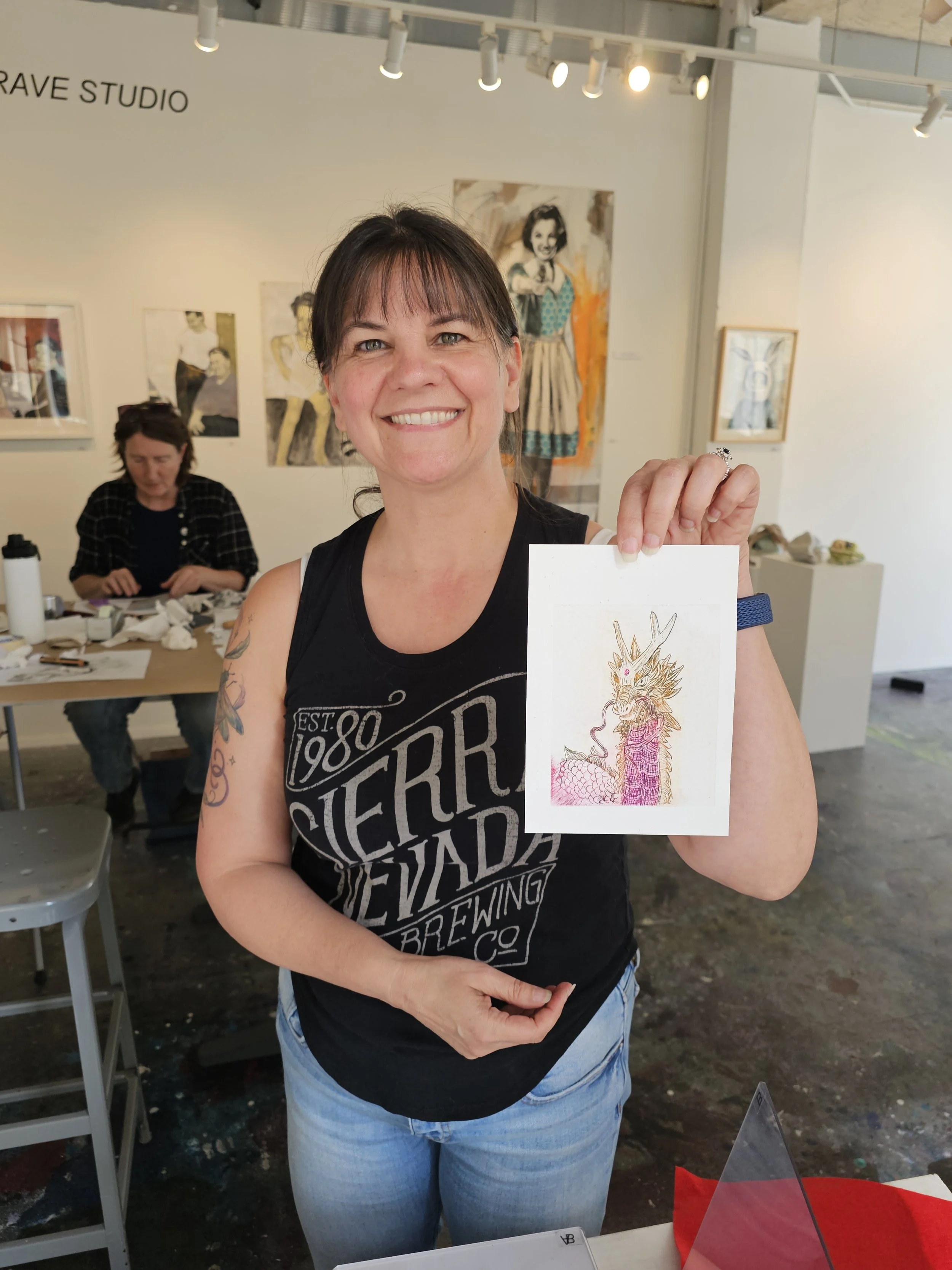 A woman smiling and holding a piece of paper with a colorful drawing of a dragon. She has dark hair, tattoos on her arms, and is wearing a black sleeveless shirt and jeans. In the background, another woman is seated at a table with art supplies, and there are paintings on the white gallery wall behind them.