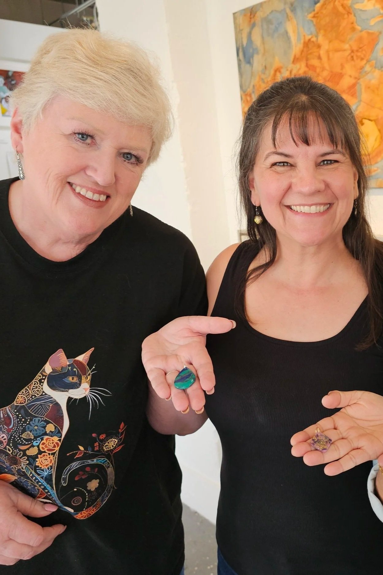 Two smiling women showing off their colorful jewelry, including rings and earrings, standing in front of an abstract orange and blue painting.
