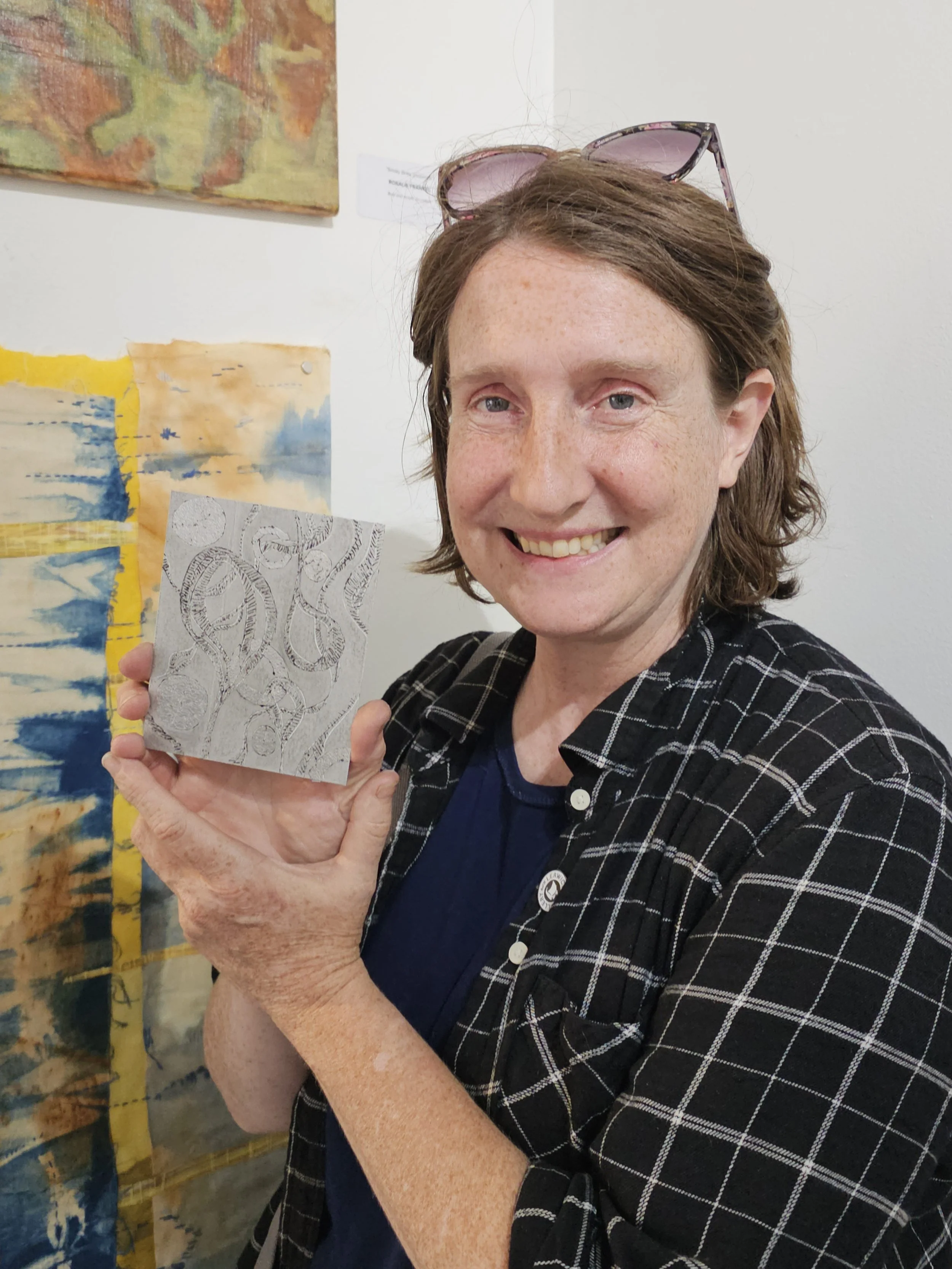 A smiling woman with short brown hair, wearing glasses on her head, holding a piece of artwork with a black and white abstract design. She is dressed in a black and white checkered shirt and a dark blue top, standing in front of colorful paintings on a white wall.