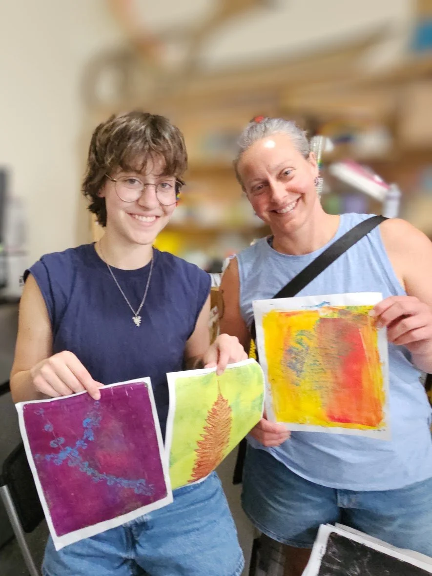 Two women smiling and holding colorful abstract paintings in an art classroom.