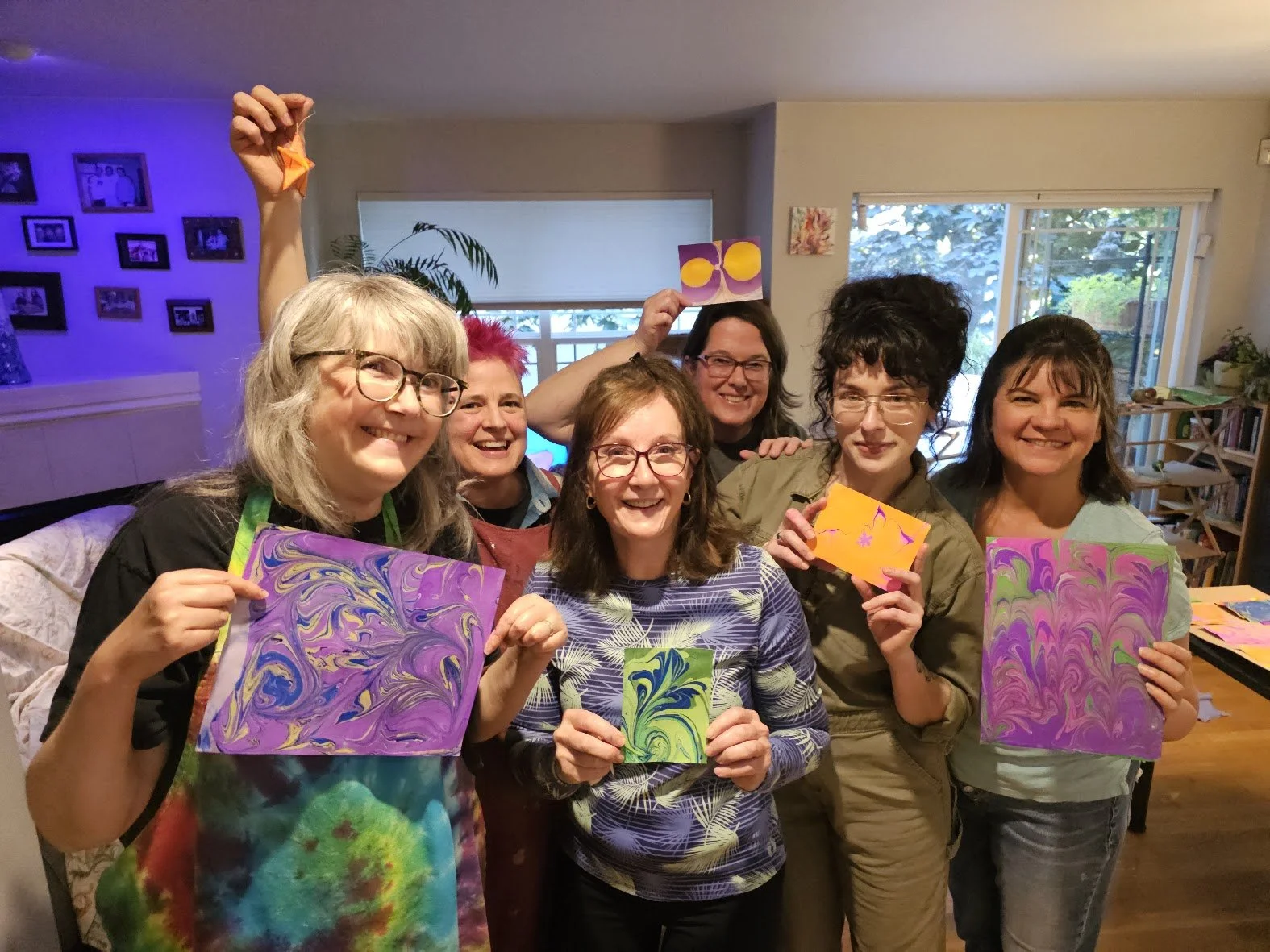 Group of women smiling and holding colorful marbled paper artworks in a cozy indoor setting with photos on the wall and large windows in the background.