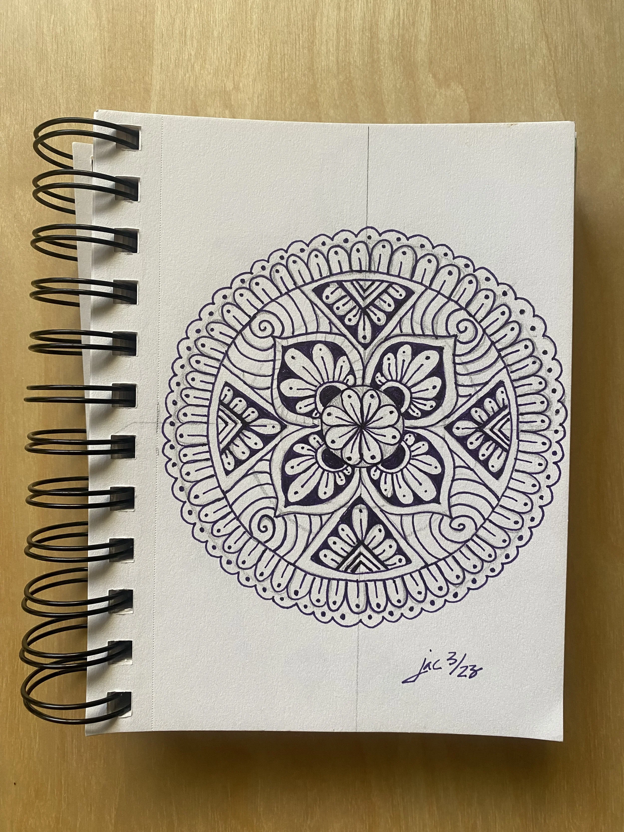 A detailed black and white mandala drawing on a spiral-bound notebook page. The mandala features intricate patterns with concentric circles, floral motifs, and geometric shapes. The page has a handwritten note at the bottom right corner that reads "Lac 3/23." The notebook is placed on a light wooden surface.