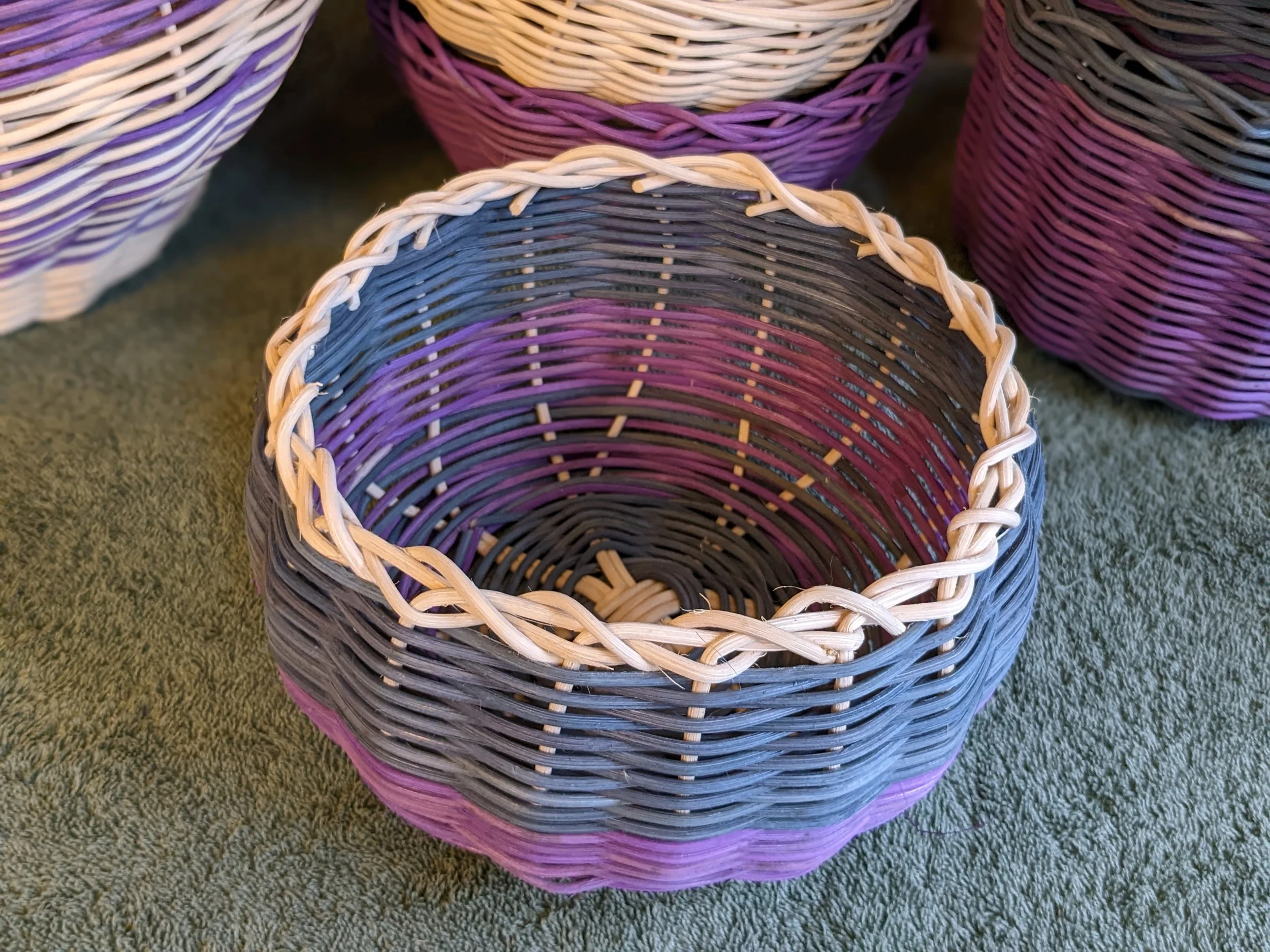 Colorful woven baskets in shades of purple, pink, gray, and cream displayed on a greenish carpet.