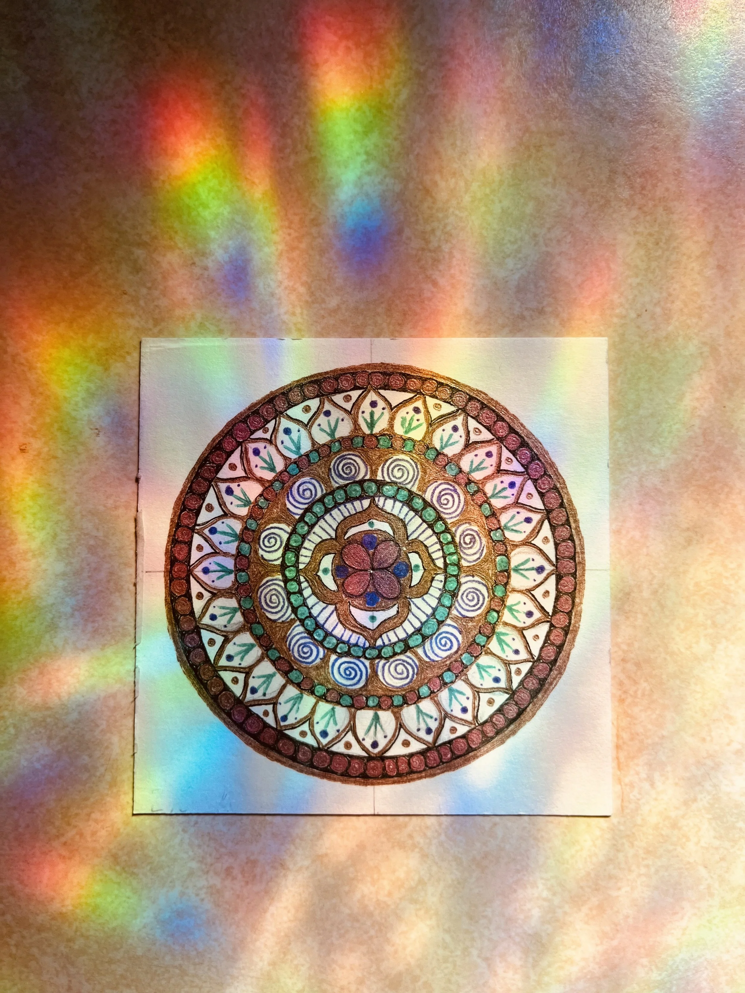Colorful, intricate mandala design with various geometric shapes and patterns on paper, with rainbow light reflections on surface.