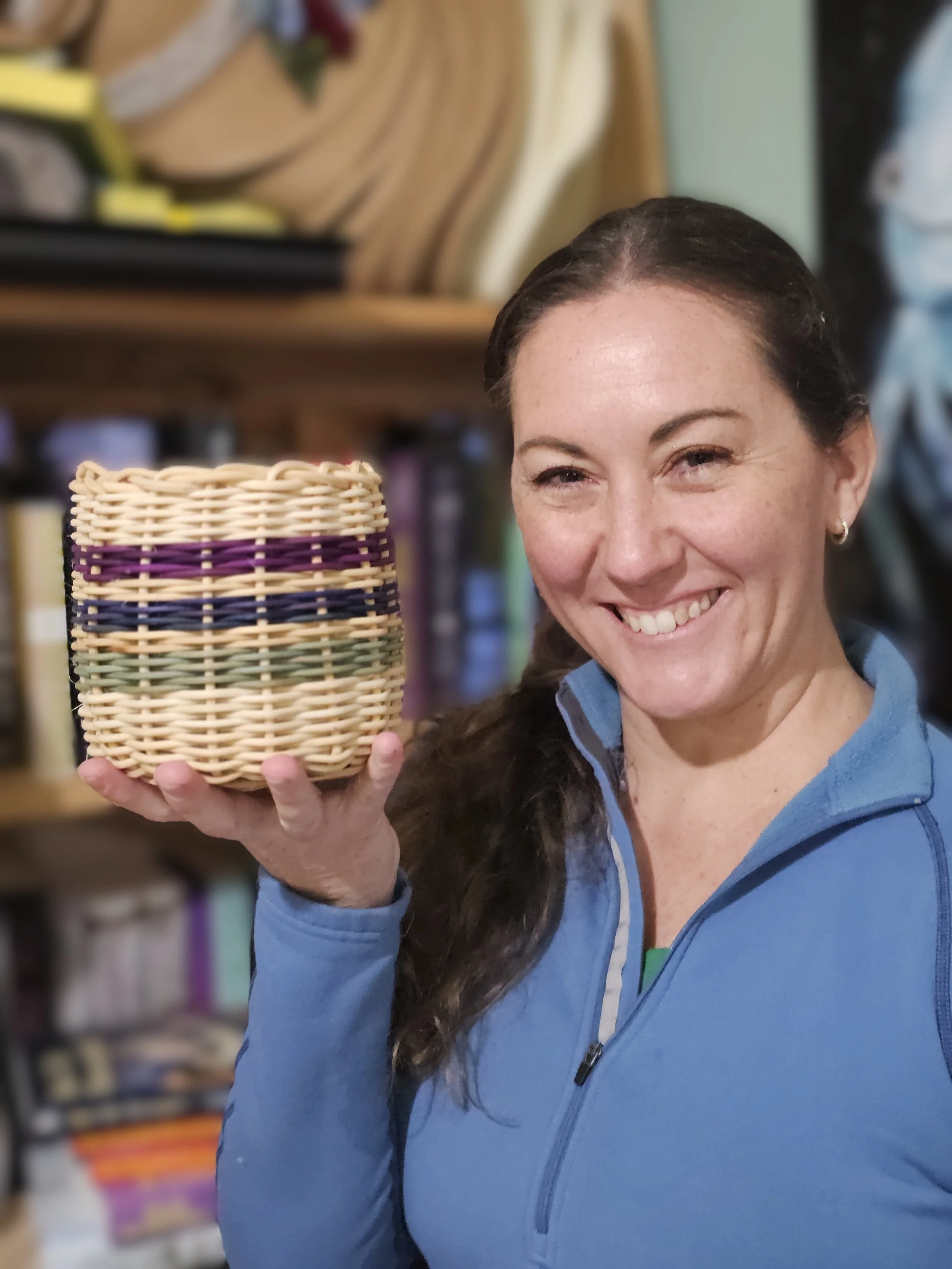 Smiling woman in a blue jacket holds a colorful woven basket in a craft room.