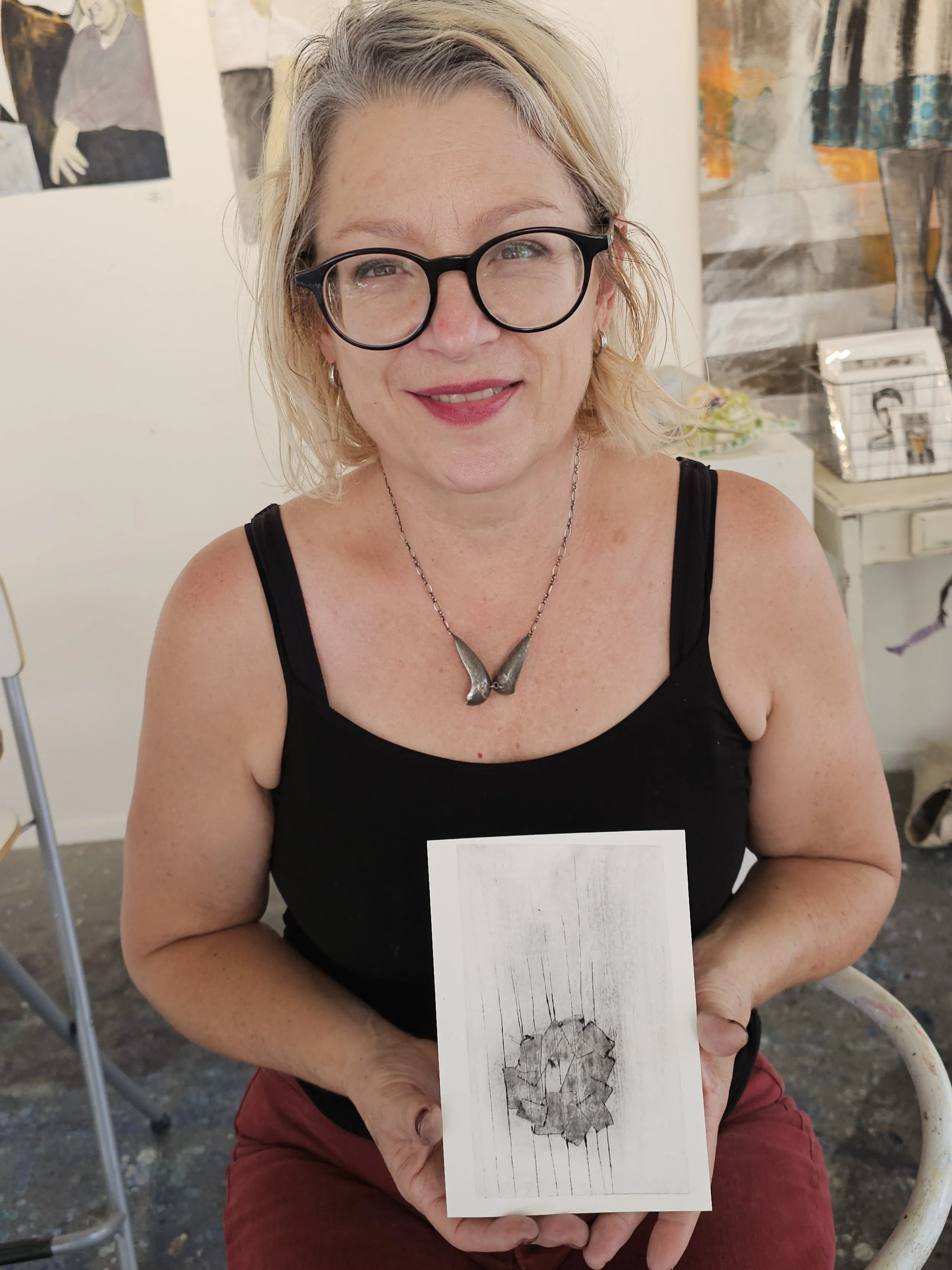 A woman with blonde hair, glasses, and a black tank top is sitting and smiling at the camera while holding a card with a cracked glass artwork in front of her. The background shows a wall with paintings and some furniture.