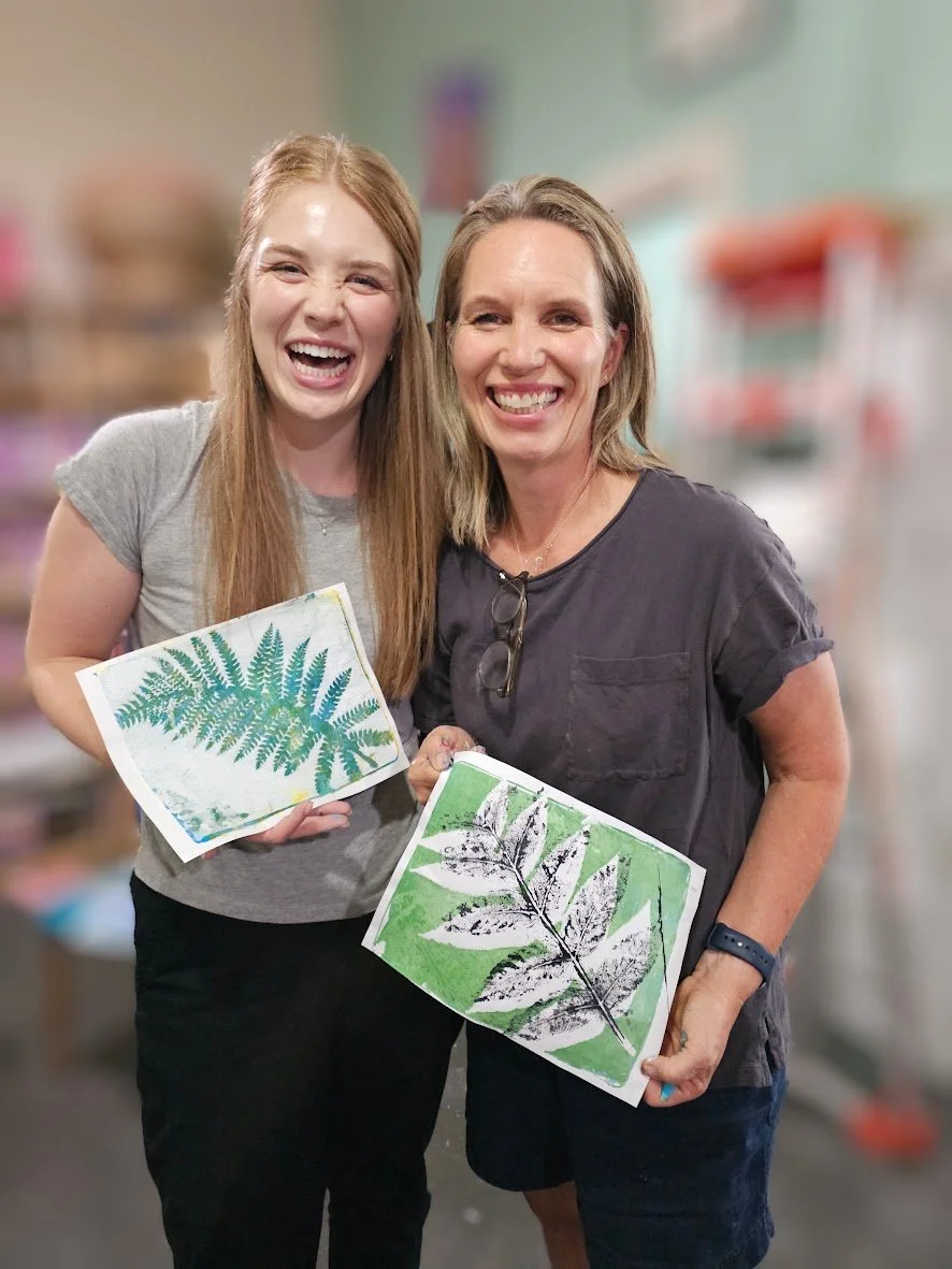 Two women smiling and holding artistic leaf prints, standing in a store or workshop.
