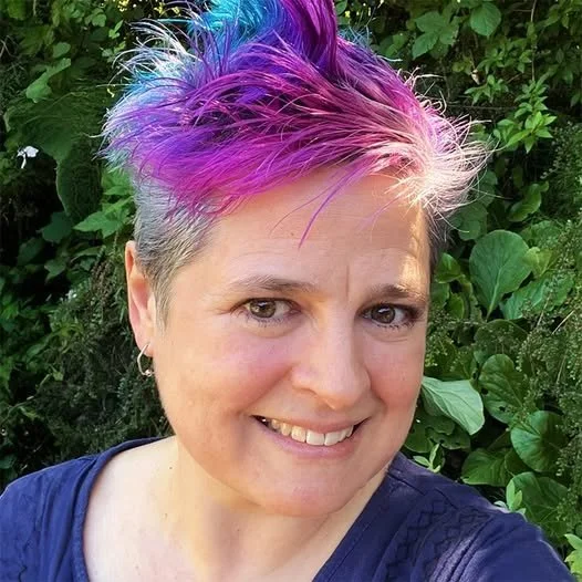 Woman with short hair dyed in shades of purple, blue, and pink, smiling outdoors with green foliage in the background.