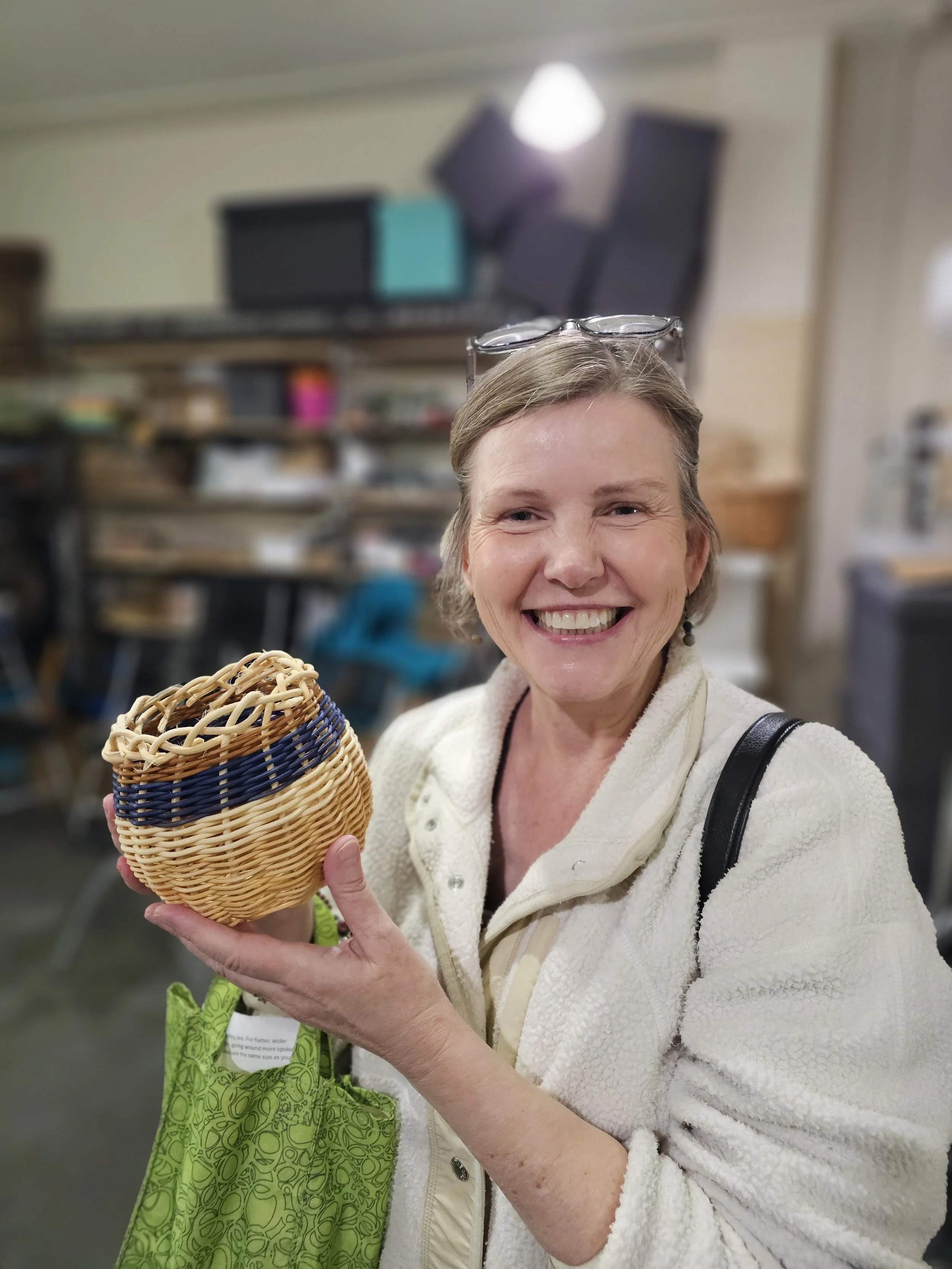 Smiling woman with glasses on her head holds a small woven basket in a shop.