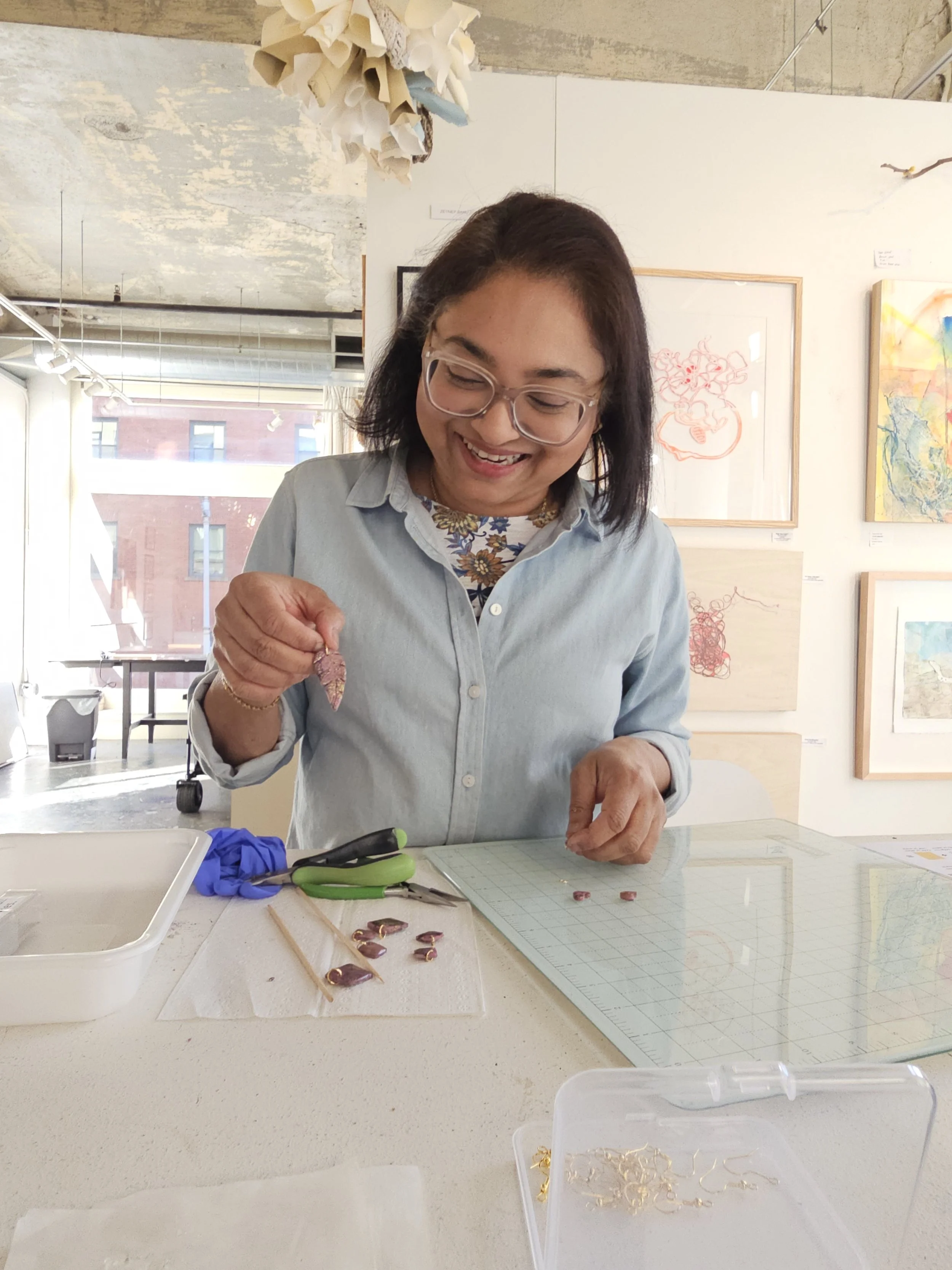 A woman with glasses and dark hair, smiling, working with jewelry tools at a table in an art gallery. She is holding a small jewelry piece, with other jewelry components laid out on the table. Artwork and frames are on the white walls behind her.