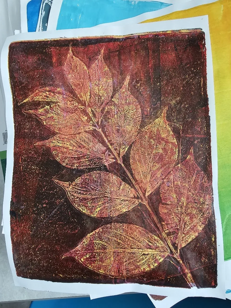 Photograph of a preserved leaf imprint with a reddish background, showing the detailed veining of multiple leaves in yellow, orange, and red hues.