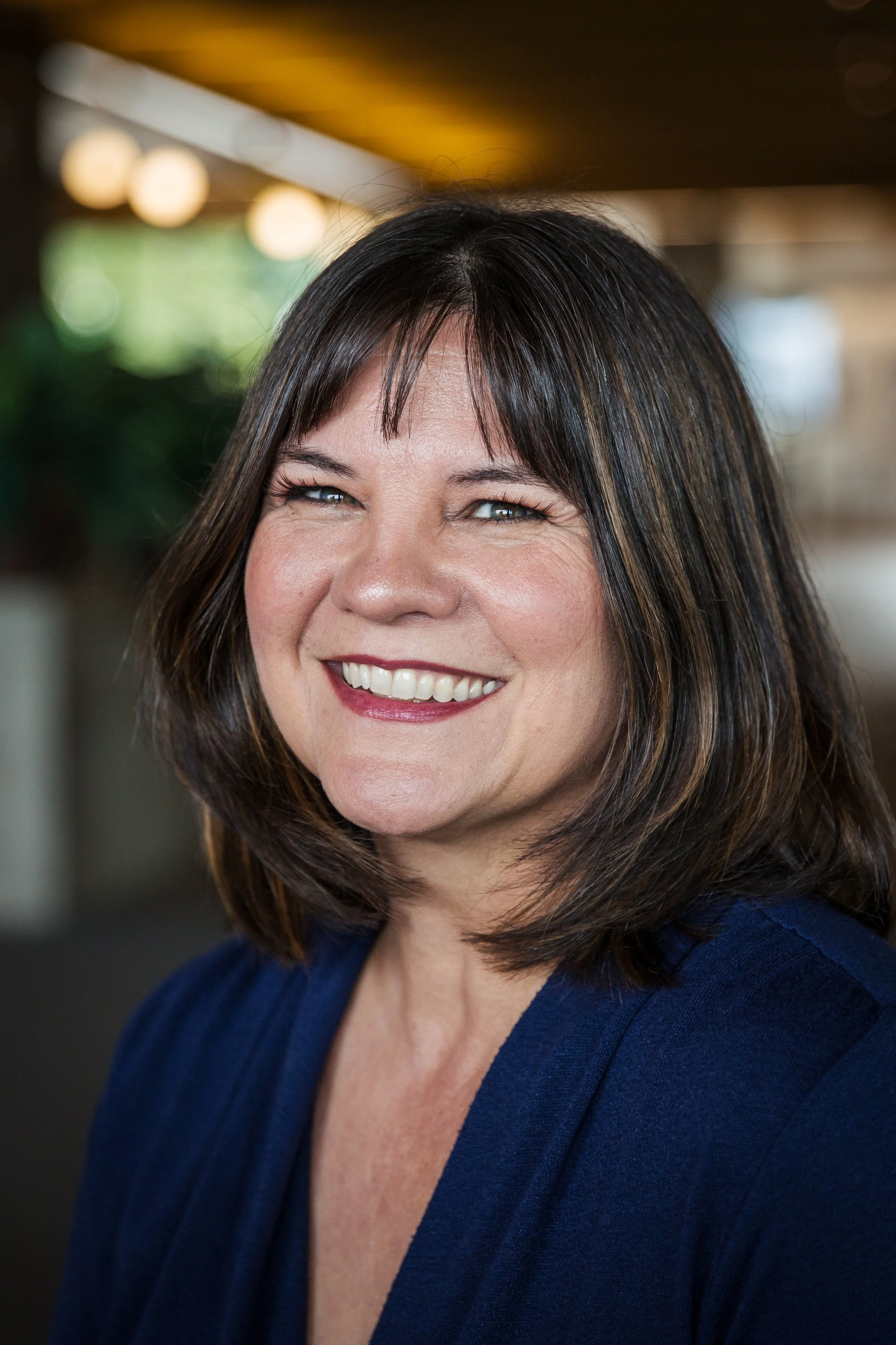 Close-up of a smiling middle-aged woman with shoulder-length dark hair, wearing a navy blue blazer, in an indoor setting with blurred lights and background.