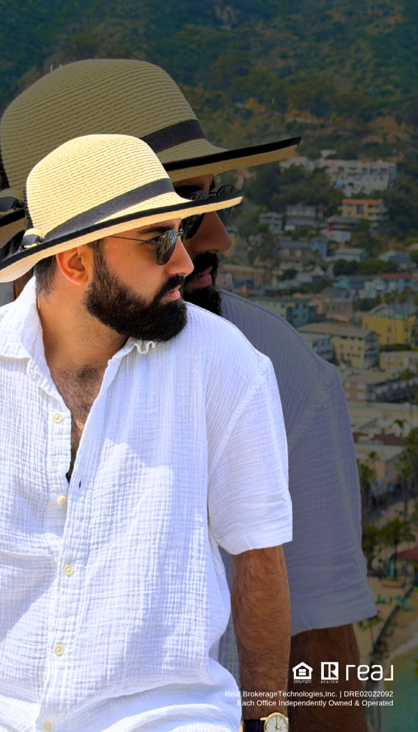 A man wearing a beige hat with a black band and sunglasses, dressed in a white button-up shirt, standing outdoors with a hillside town in the background.