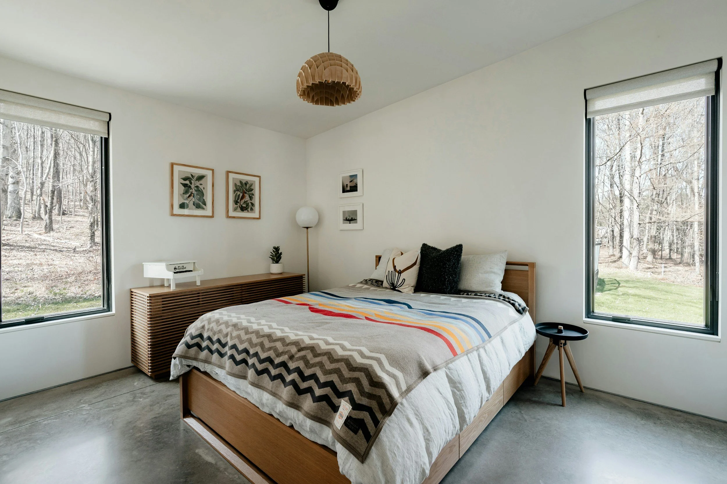 A minimalist bedroom with large windows showing a wooded outdoor view, a bed with a colorful striped blanket, framed artwork on the white wall, a small table on black hairpin legs, and a wooden sideboard with a potted plant and a white lamp.