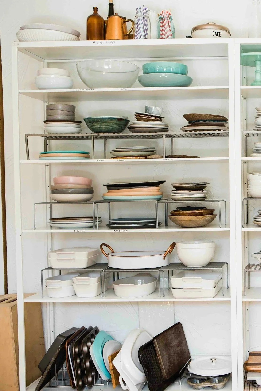 White kitchen shelves filled with various dishes, bowls, and cookware, mainly in neutral tones with some pastel and earthy accents, organized on a white shelf.