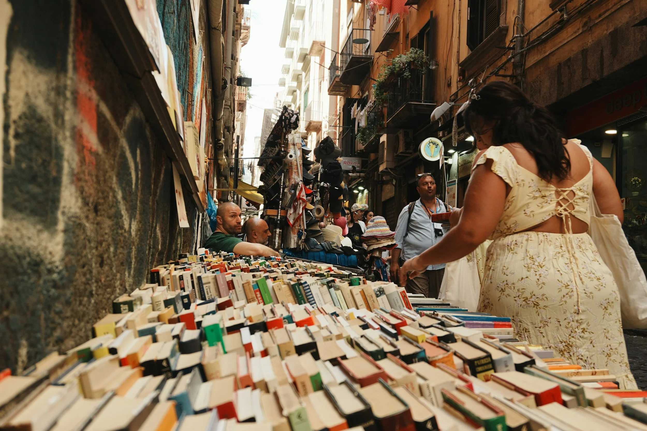 Street market with an outdoor book sale on a narrow alley, books laid out on tables, and people browsing.