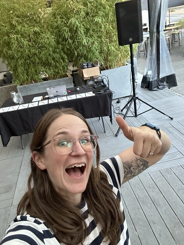 A woman with long brown hair and glasses taking a selfie at an outdoor event with a happy expression, pointing upward. In the background, there is a table with electronic equipment, a large speaker on a stand, and some greenery.
