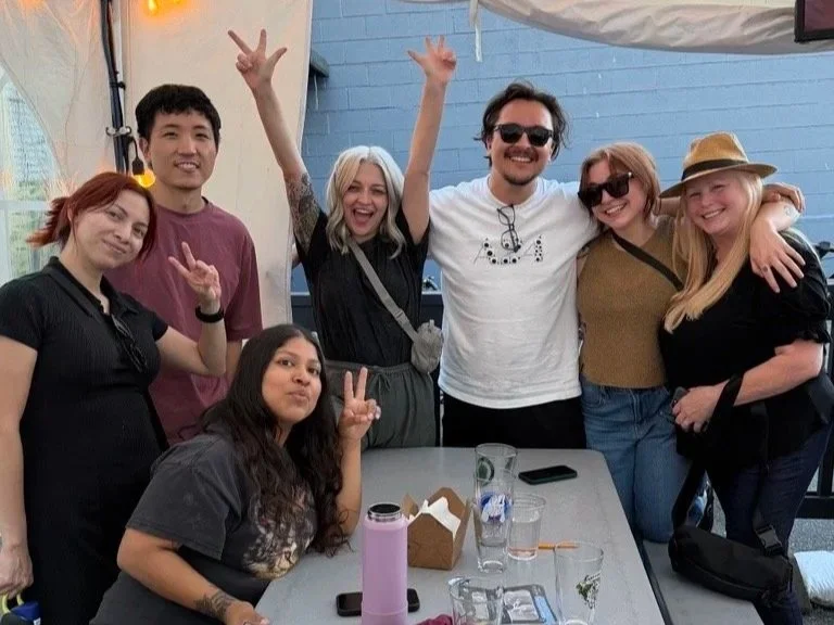 Group of seven diverse friends smiling and making peace signs during Trivia Night, standing around a table with drinks and food.