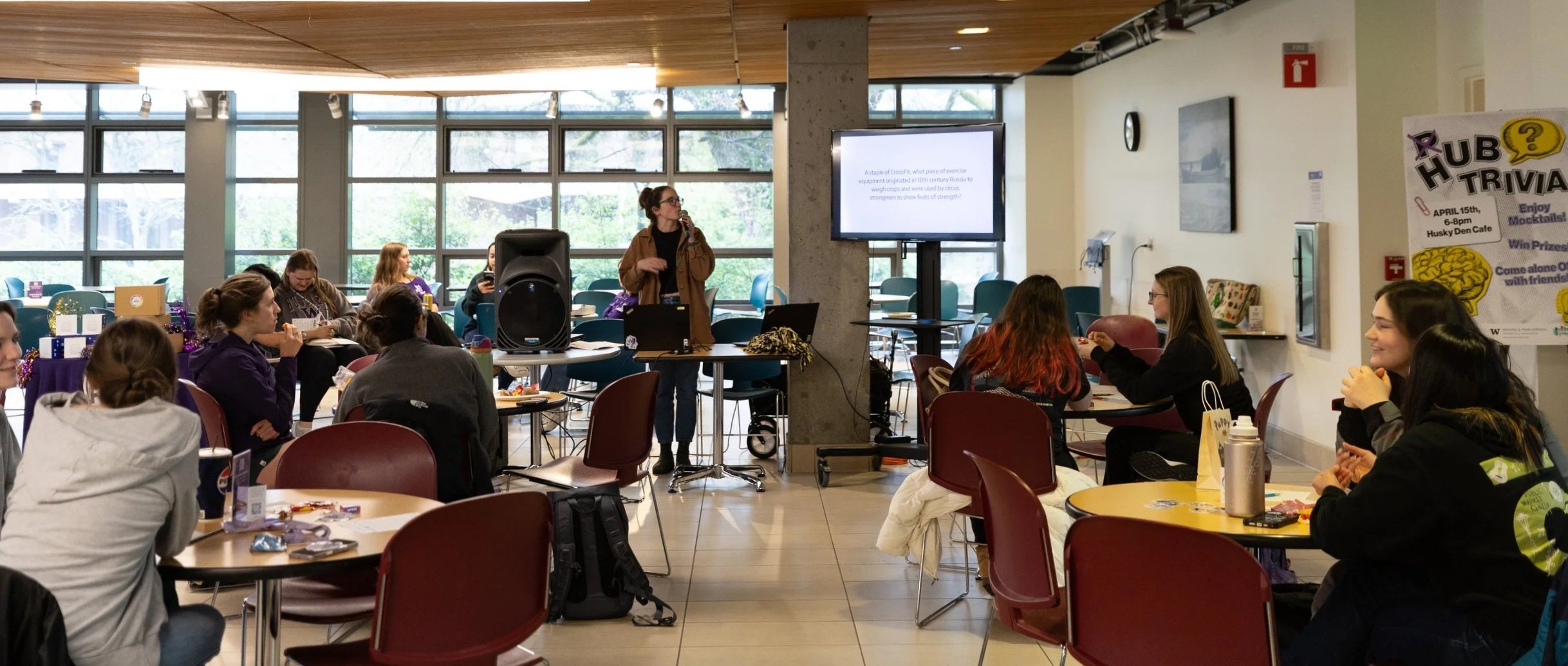 A woman in glasses and a brown jacket hosts HUB Trivia Night to a group of students seated at tables in the UW student union